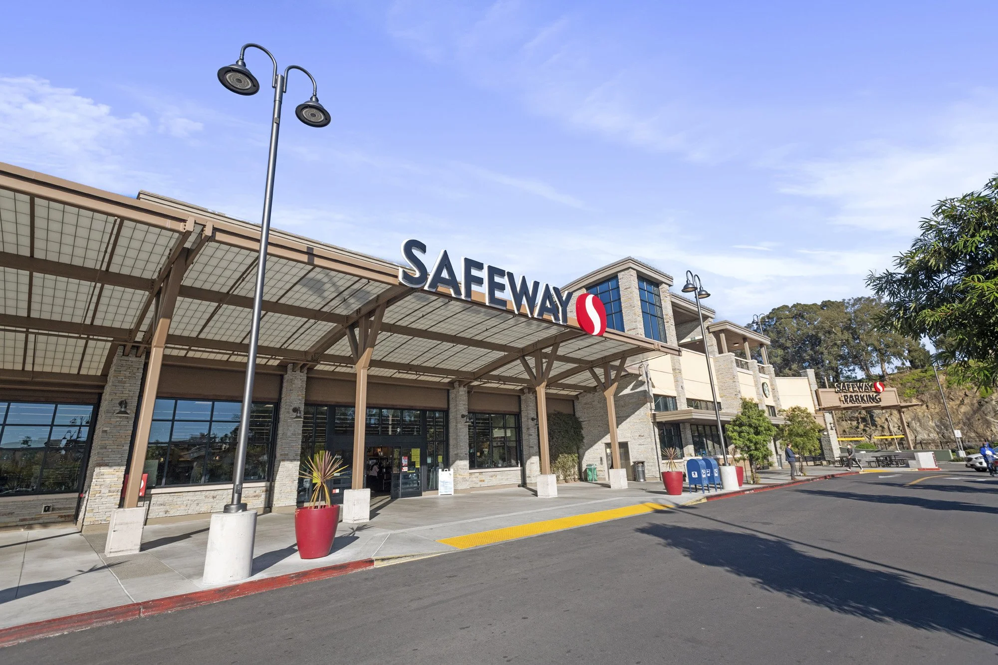Exterior of a Safeway supermarket with parking lot, signage, two street lamps, and a clear blue sky.