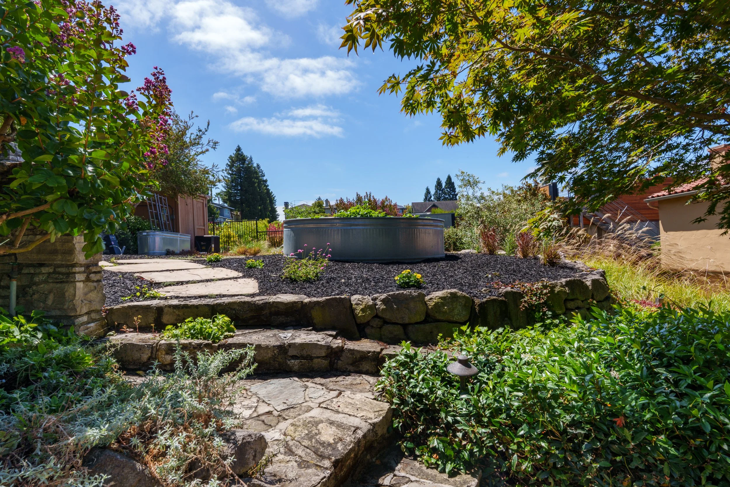 A backyard garden with stone steps leading up to a landscaped flower bed. The garden has lush greenery, flowering bushes, and a large tree with overhanging branches. In the background, there is a wooden shed and a fenced yard under a bright blue sky 