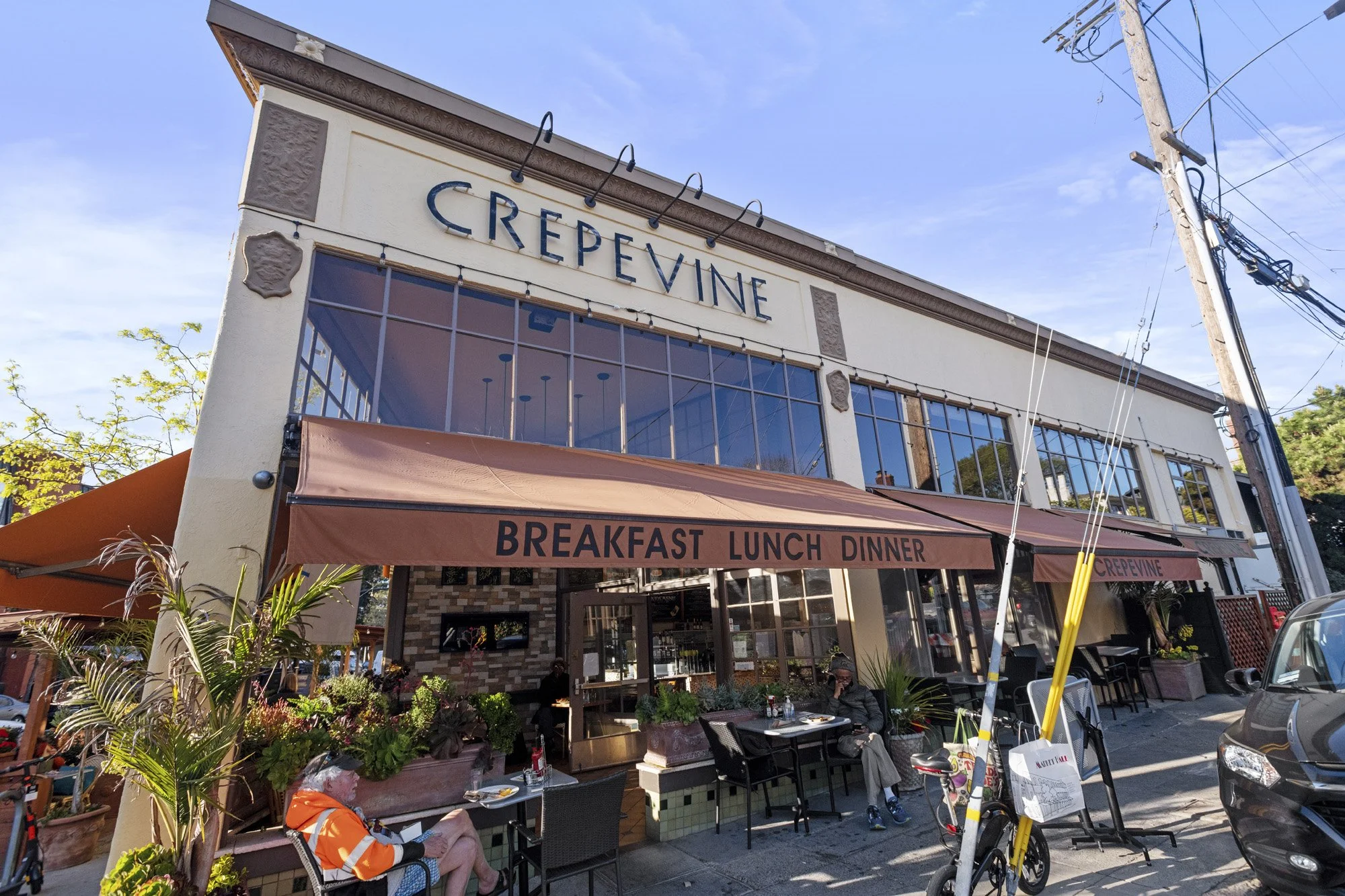 Exterior of a restaurant named Crepevine with large windows and decorative stone accents. Outdoor seating area with tables and chairs, some people dining, and an orange awning that says breakfast, lunch, dinner. Overhead power lines and a utility pol