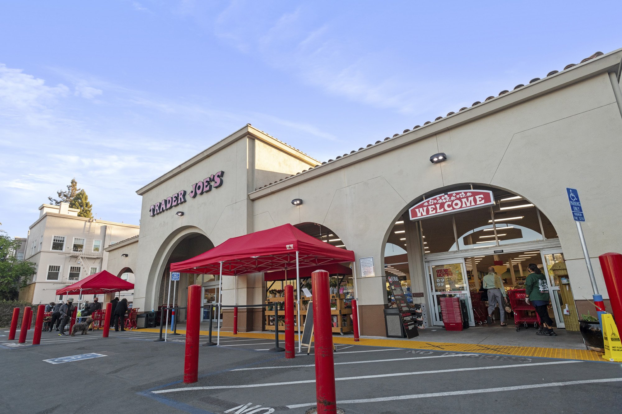 Exterior view of Trader Joe's grocery store with red tents set up outside, customers shopping, and a walk-in entrance with a welcoming sign. The building has a beige stucco facade under a blue sky with some clouds.