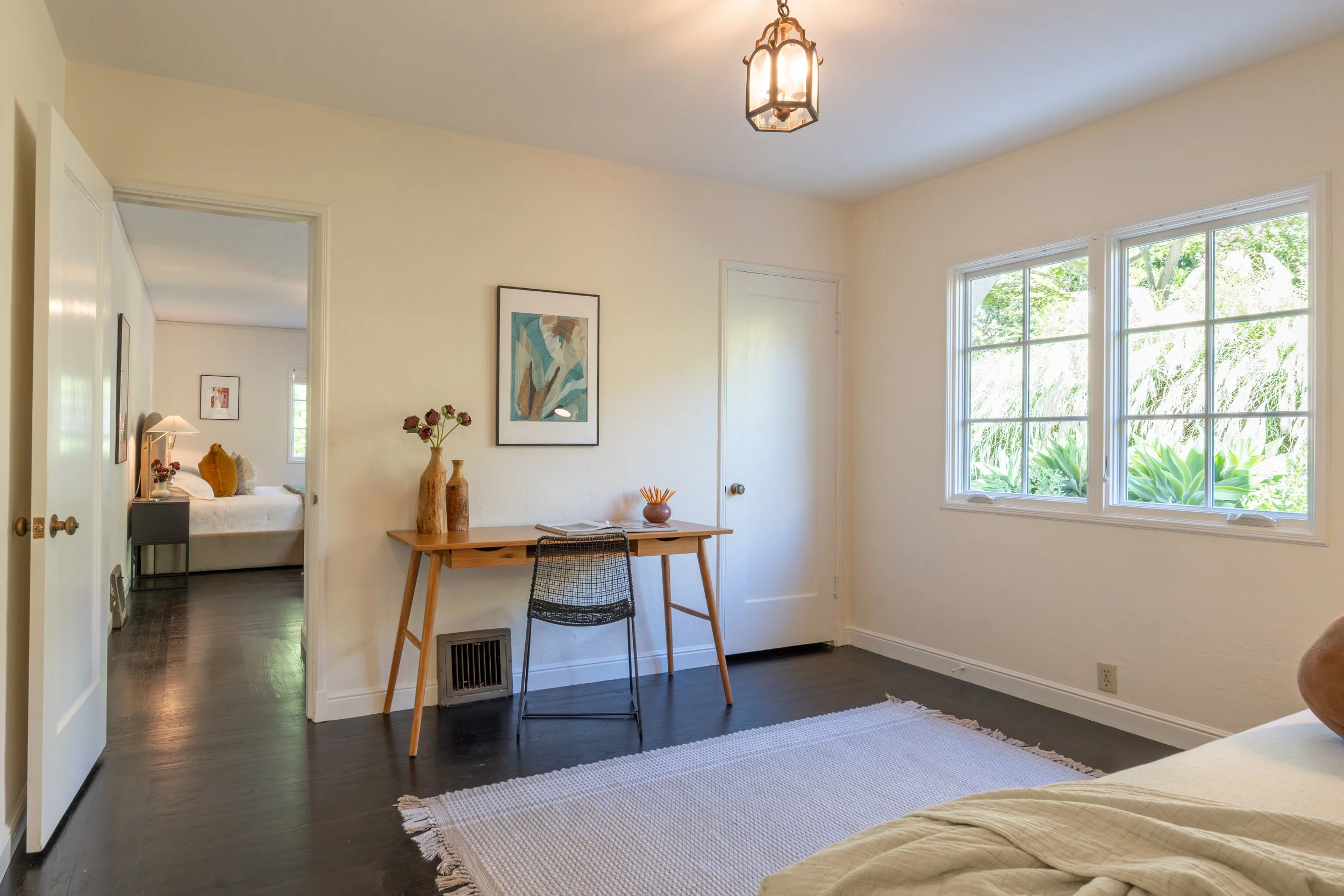 Living room with large window, wooden dining table, black chair, framed artwork, and decorative vases. Bedroom visible through doorway with bed, pillows, and lamp.