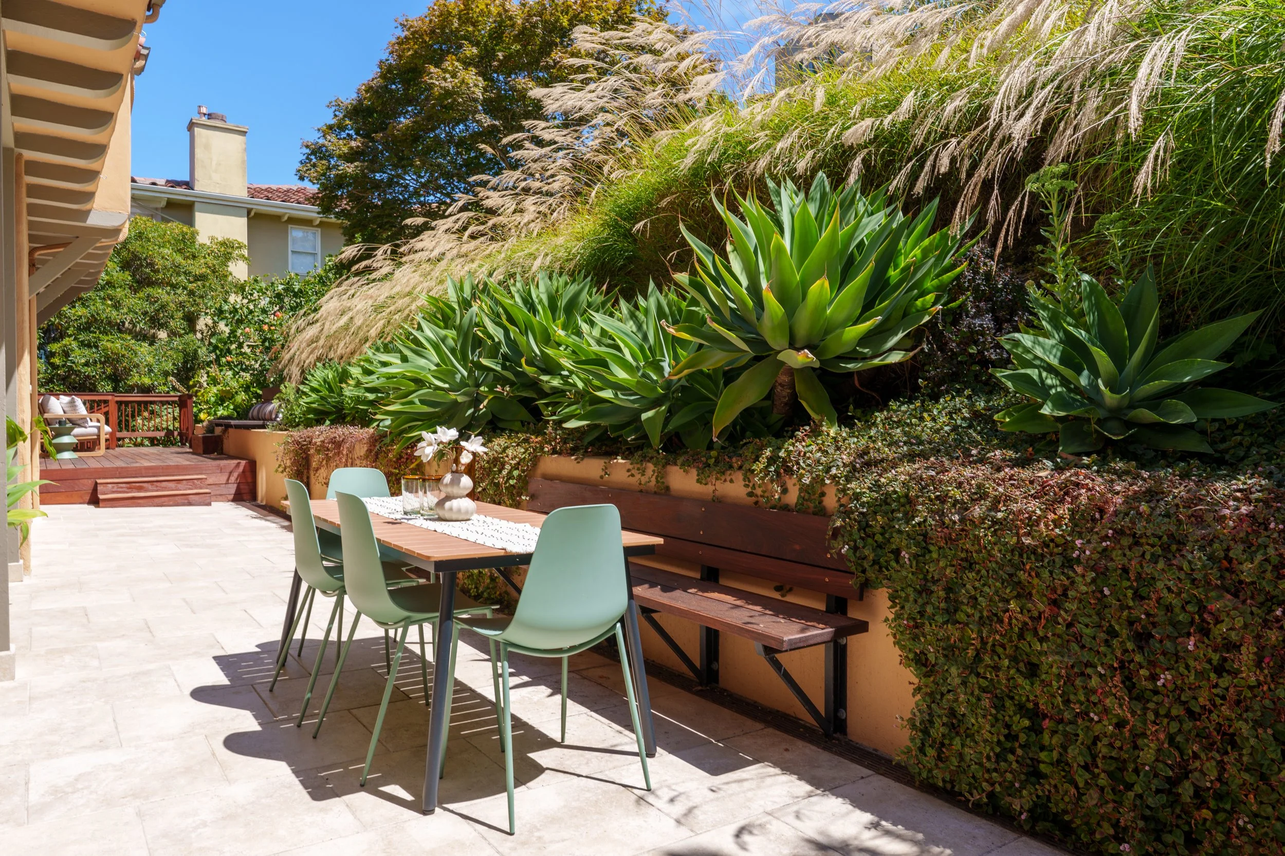 An outdoor patio area with a wooden dining table, mint green chairs, white flowers in a vase, and surrounding lush greenery including large succulent plants and tall ornamental grasses, with a house in the background under a clear blue sky.