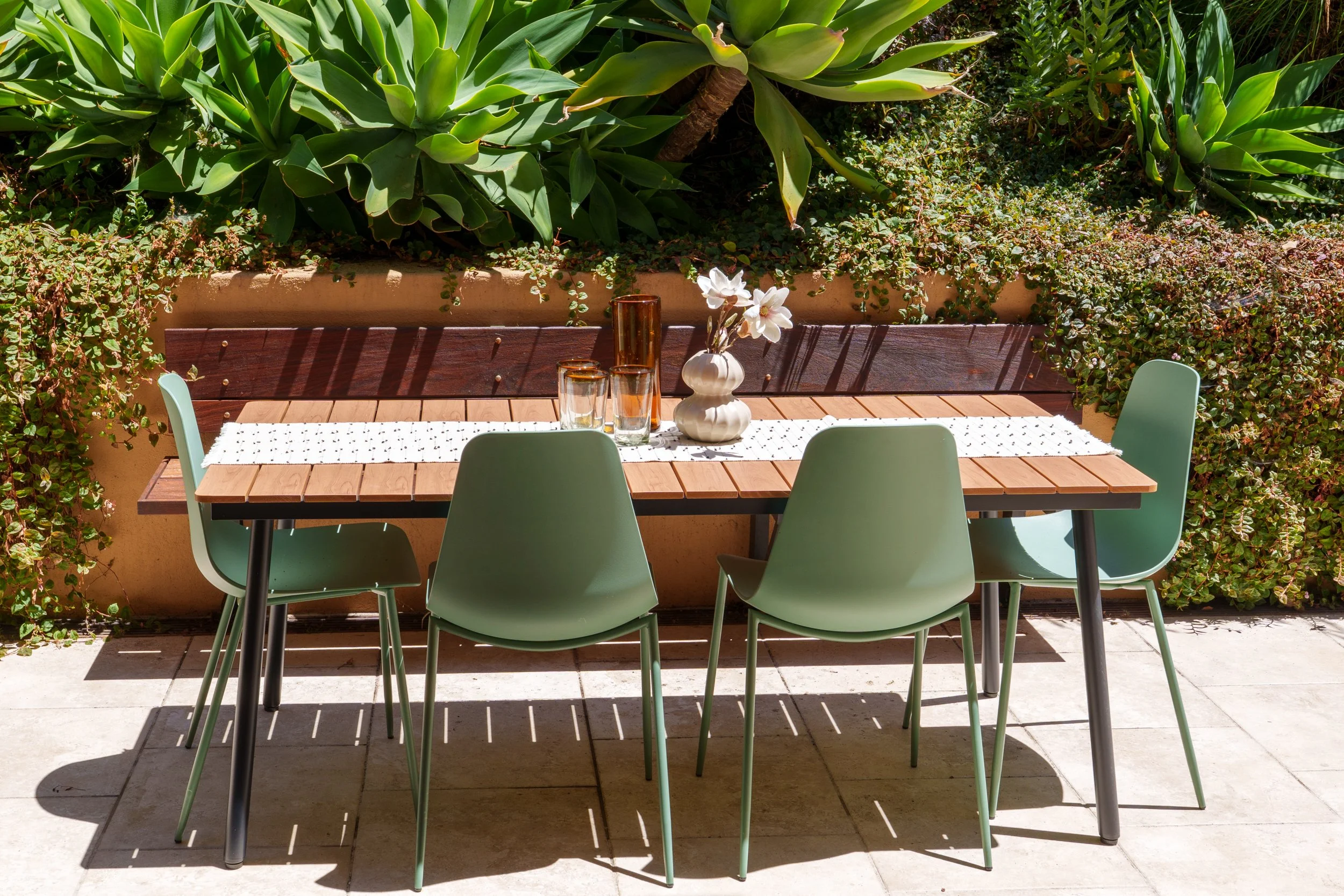 Outdoor patio dining area with a wooden table, four pastel green chairs, decorative vases with white flowers, and lush green plants in the background.