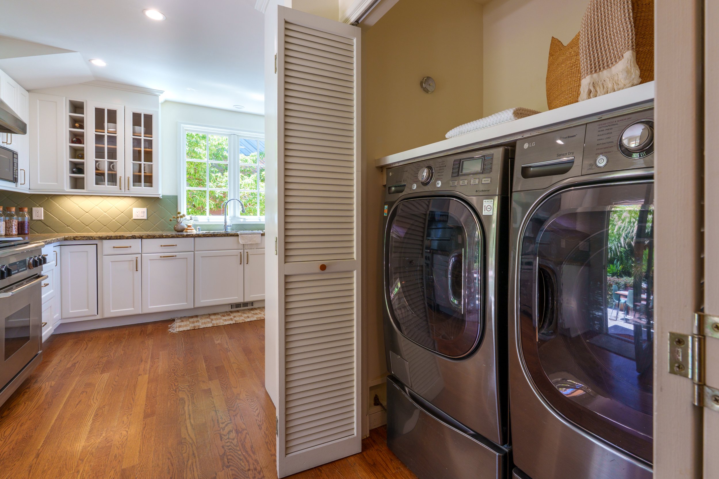 A laundry area with a front-loading washer and dryer, a white cabinet with glass doors, a window with a view of trees, and a countertop with jars.