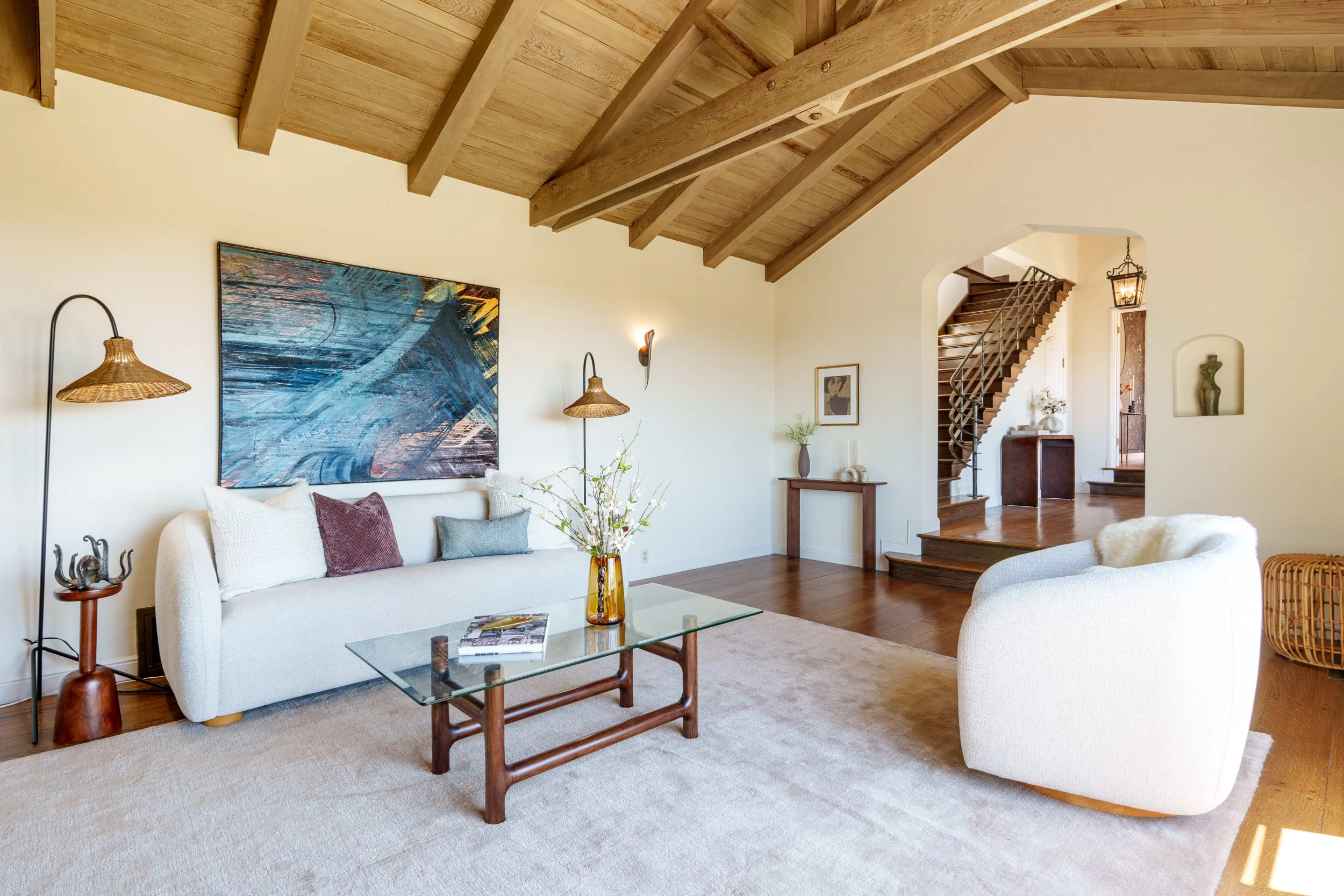 A living room with white sofas, a glass coffee table, and wooden accents. A large abstract painting hangs on the wall, and there are wicker and wooden decor items. There's a staircase with wooden steps and a railing leading to an upper level. The ceiling has exposed wooden beams.