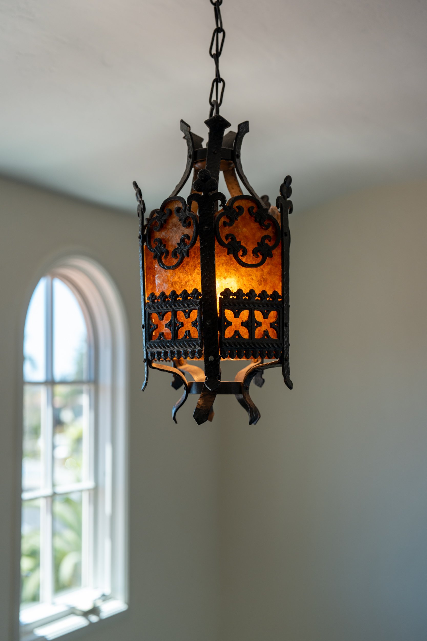 A decorative hanging lantern with intricate black metalwork and warm amber glass panels, suspended by a chain from the ceiling. In the background, a window with white trim and a view of green foliage outside.