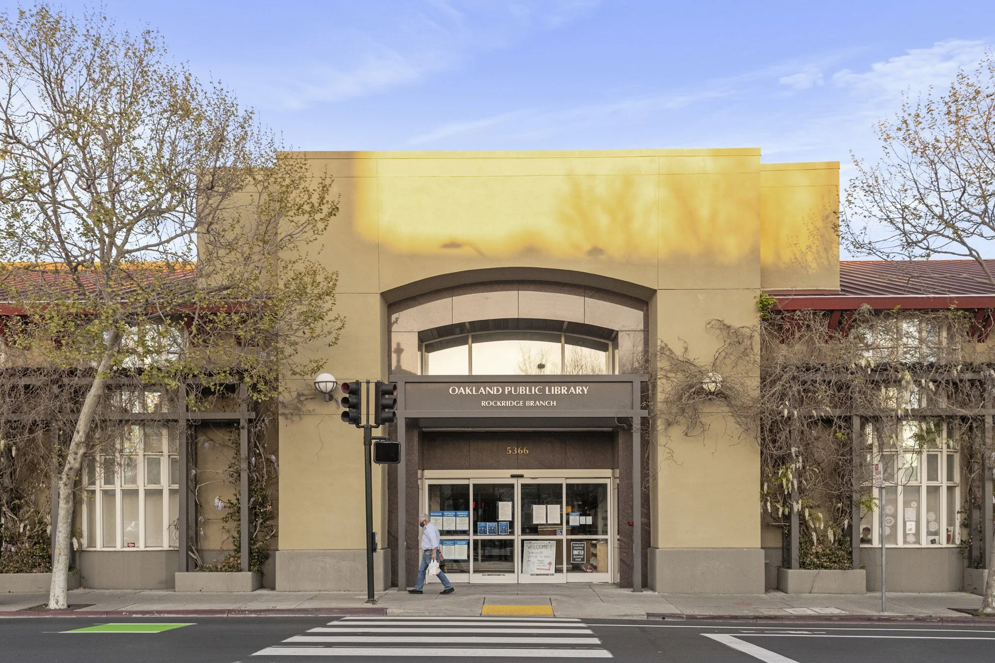 Exterior view of Oakland Public Library Rockridge Branch with a sign above the entrance and pedestrians walking outside, trees on either side, traffic lights, and a crosswalk in the foreground.