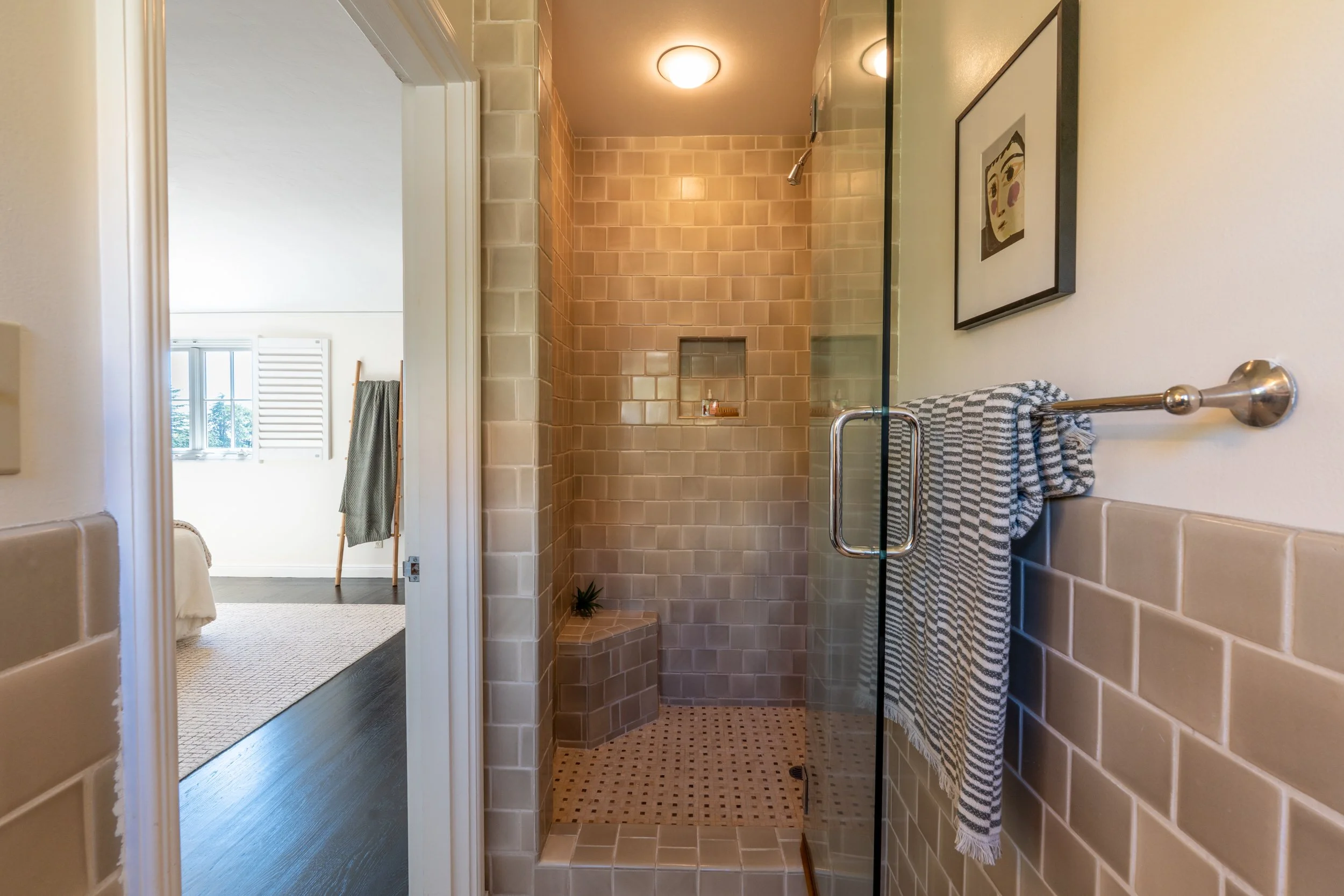 A bathroom shower with beige square tiles, a small built-in shelf, and a glass door with a towel hanging on a bar outside the shower.