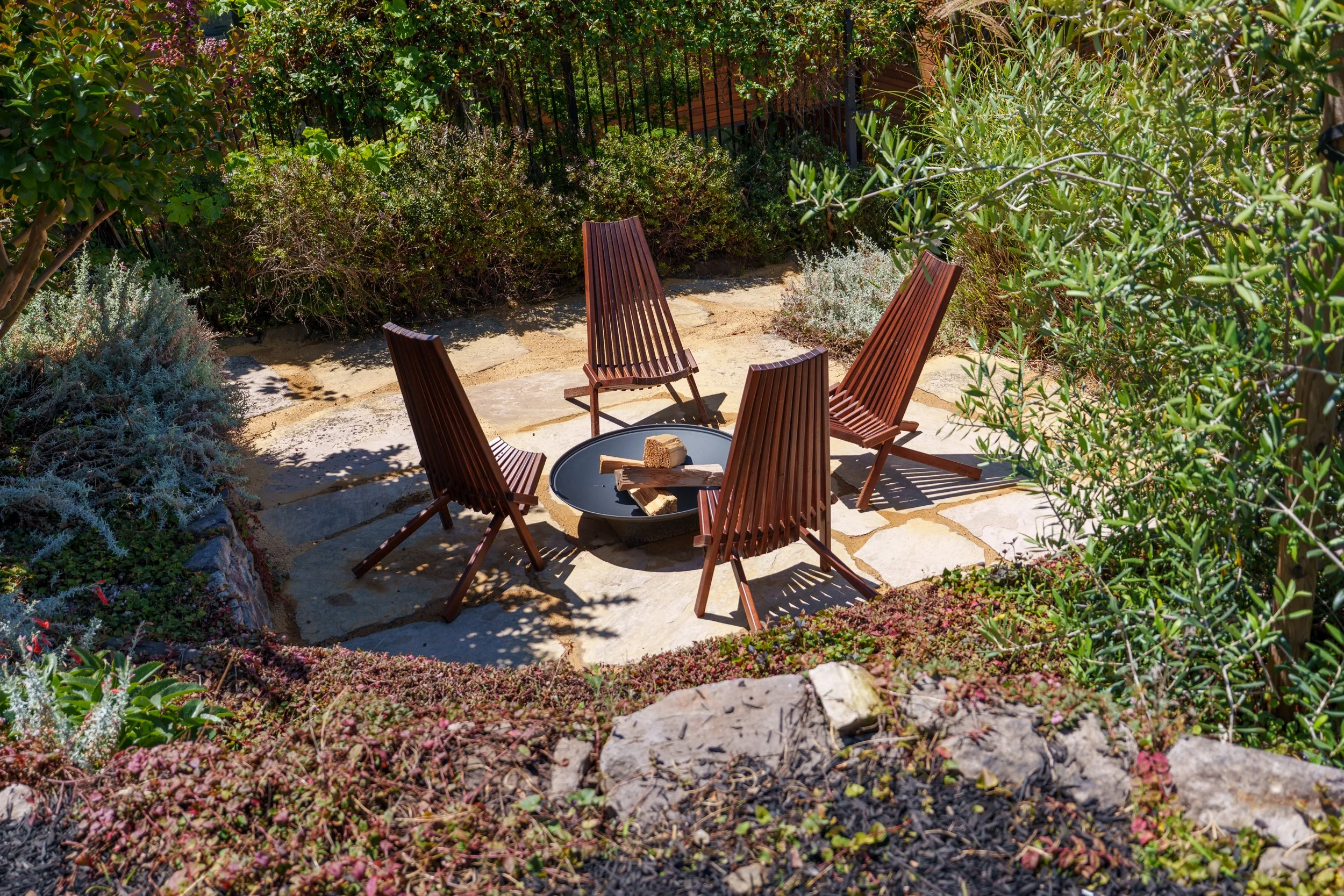 A backyard garden patio with five wooden chairs arranged around a fire pit filled with wood. The area is surrounded by lush green shrubs and plants, with a stone pathway leading to the seating area.