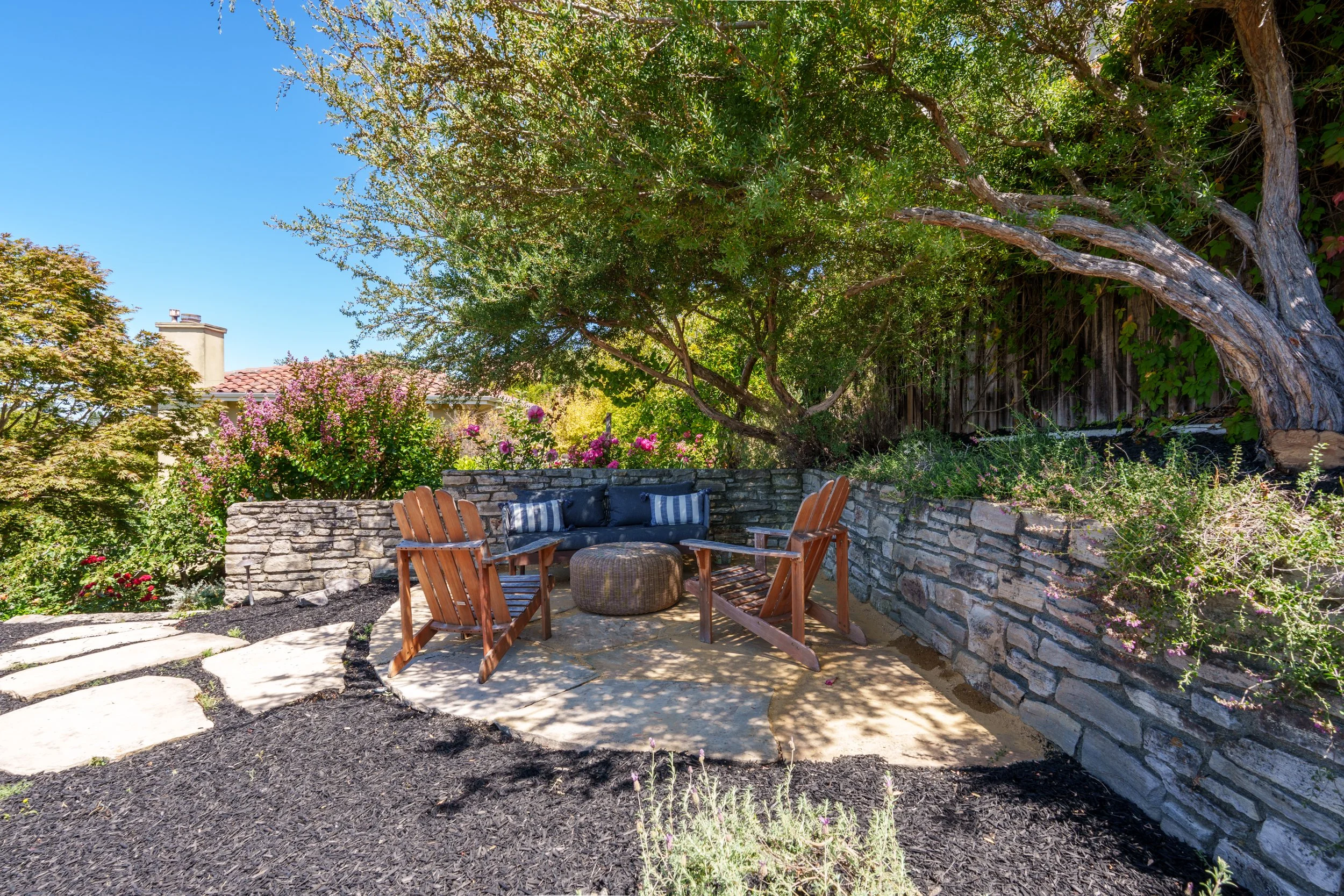 A backyard patio with wooden chairs and wicker ottoman, surrounded by lush green plants, colorful flowers, and a stone wall under a large tree, with a house partially visible in the background.