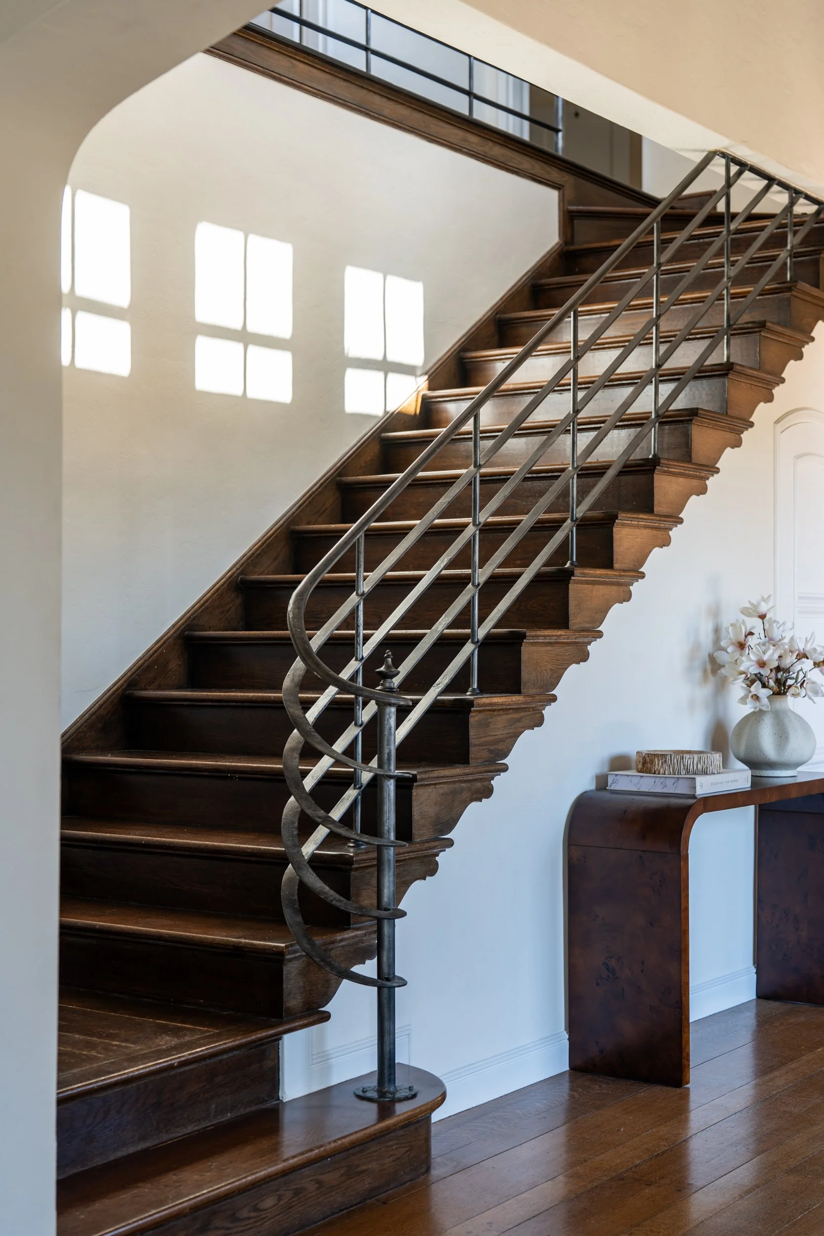 Wooden staircase with metal railing casting shadows on the wall, with a small table holding a vase with flowers nearby.