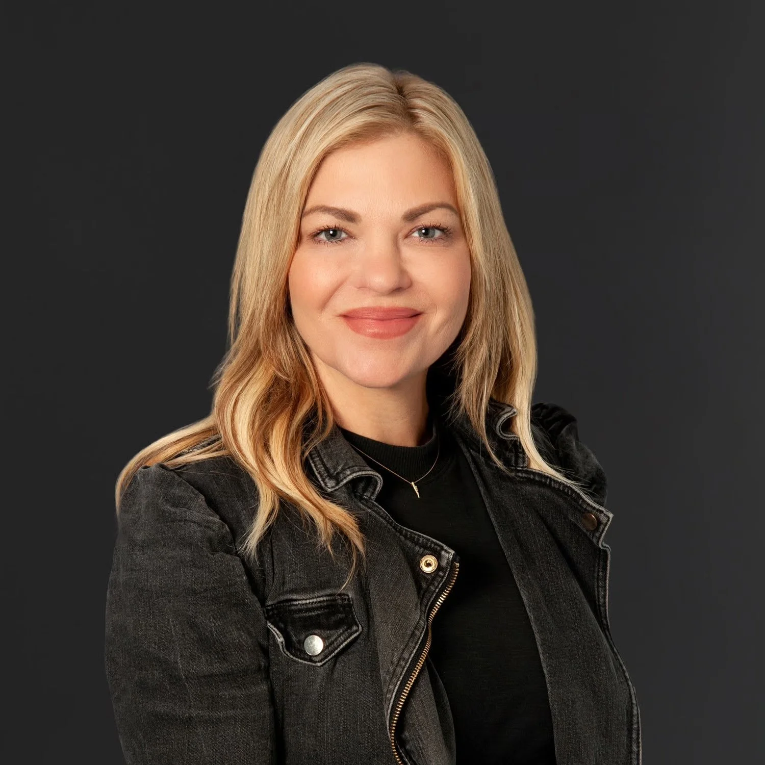 A woman with blonde hair wearing a black denim jacket and a black top, smiling with crossed arms against a dark gray background.