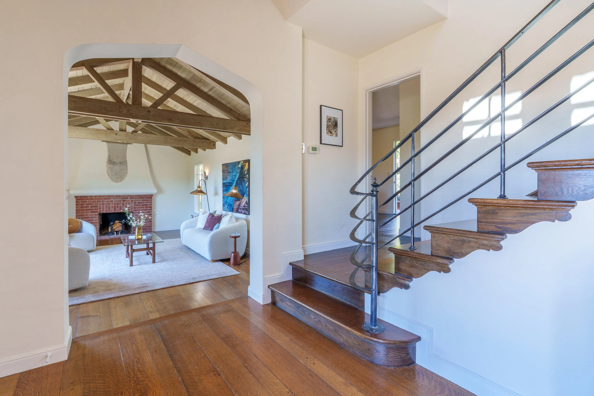 Interior view of a house showing a curved wooden staircase with a metal railing, leading to an upper floor. The lower level has a living room with white sofas, a brick fireplace, and artwork on the walls. Exposed wooden beams are visible on the ceili