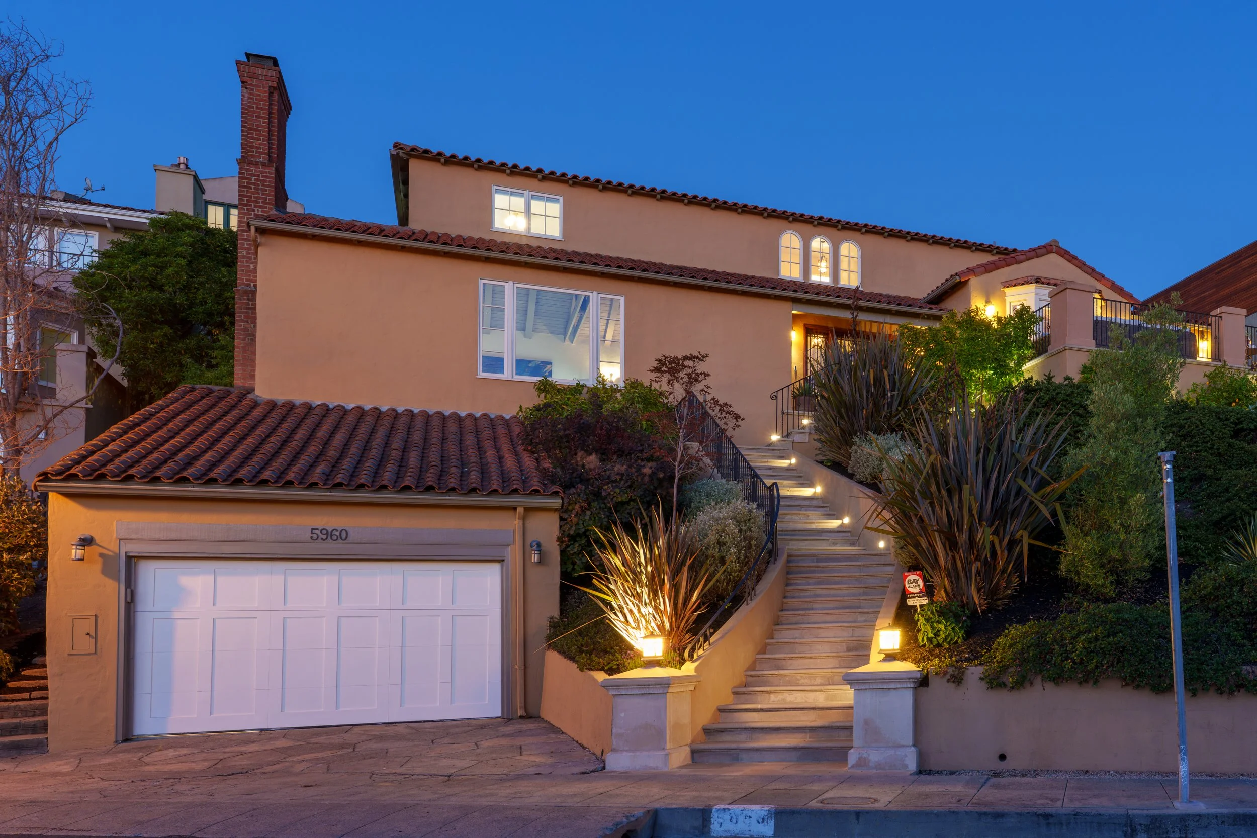A multi-story house with beige stucco walls and a red-tile roof, outdoor stairs with lighting leading to a second-floor balcony, lush greenery, and a garage door at the front.