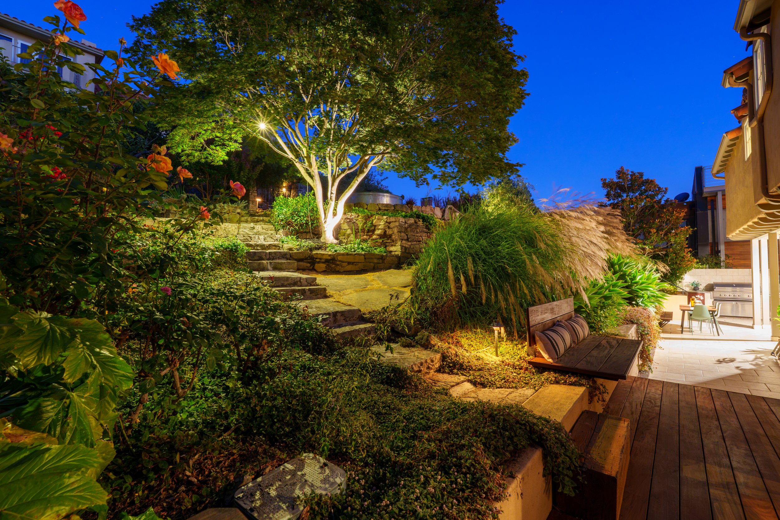 A backyard garden at dusk with a large illuminated tree, stone steps, lush plants, and outdoor furniture on a wooden patio.