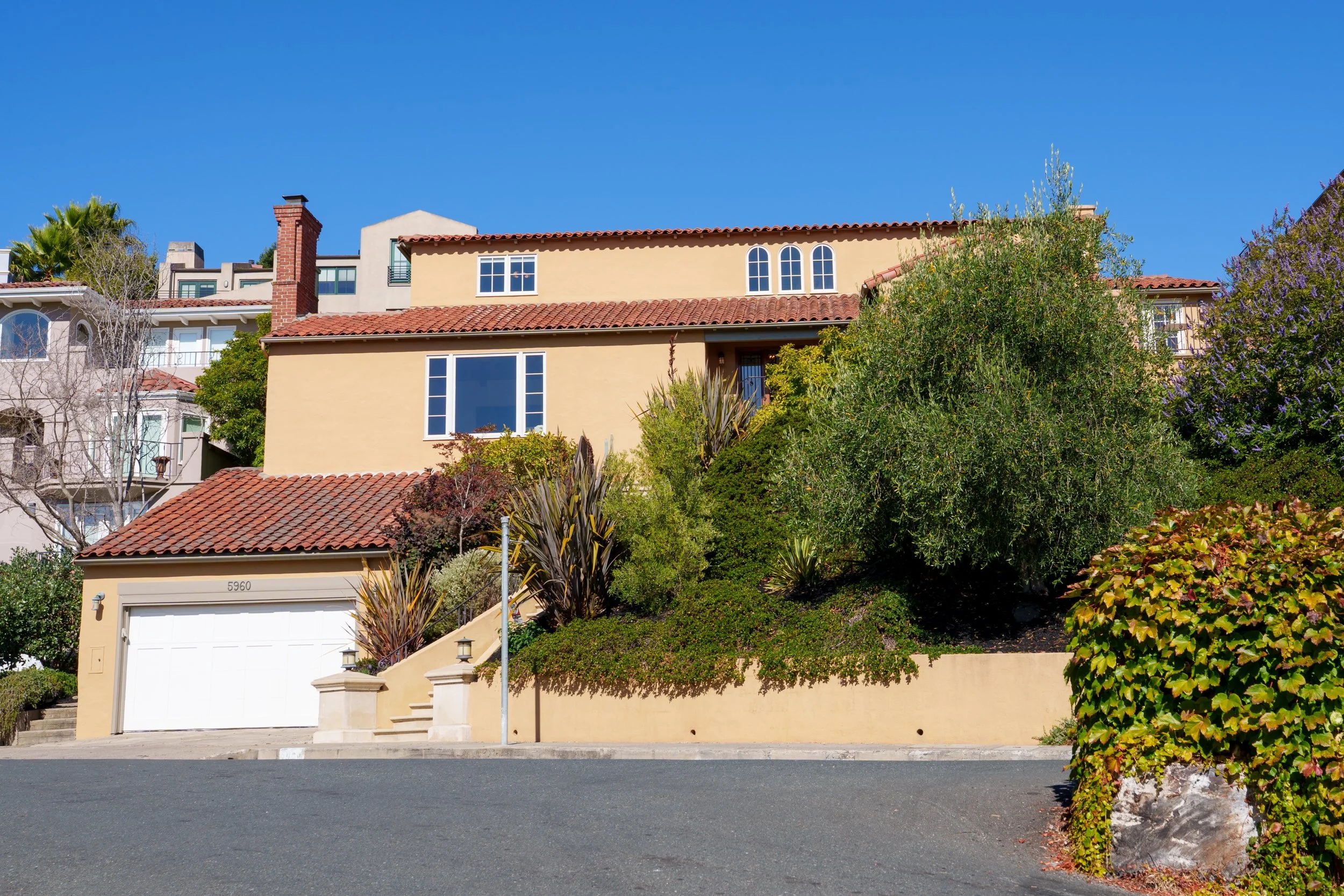 A multi-story house with a yellow exterior, red-tiled roof, and various windows, surrounded by bushes and trees under a clear blue sky.