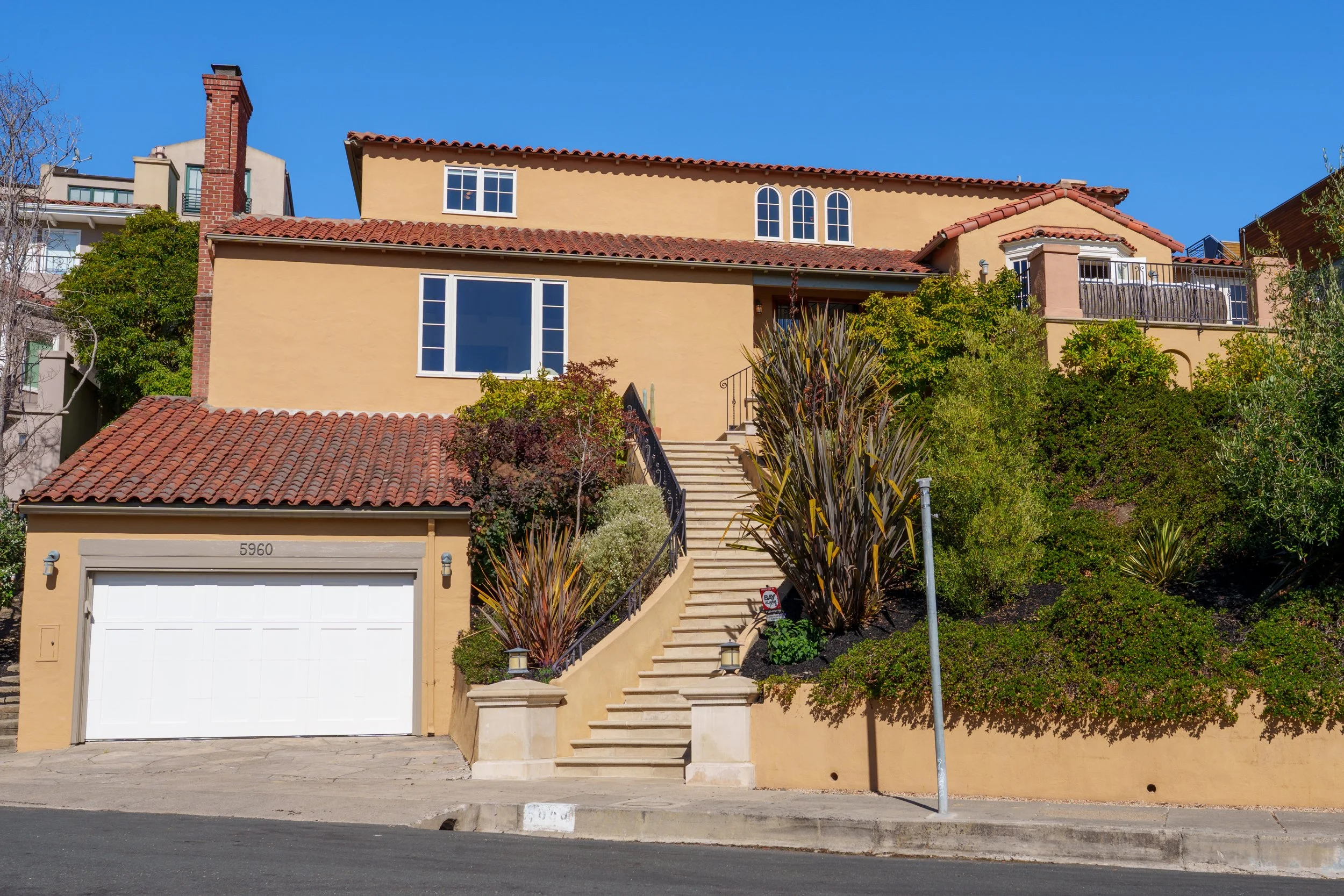 A beige multi-story house with red tiled roof, front stairs, and a garage door, surrounded by bushes and trees, under a clear blue sky.