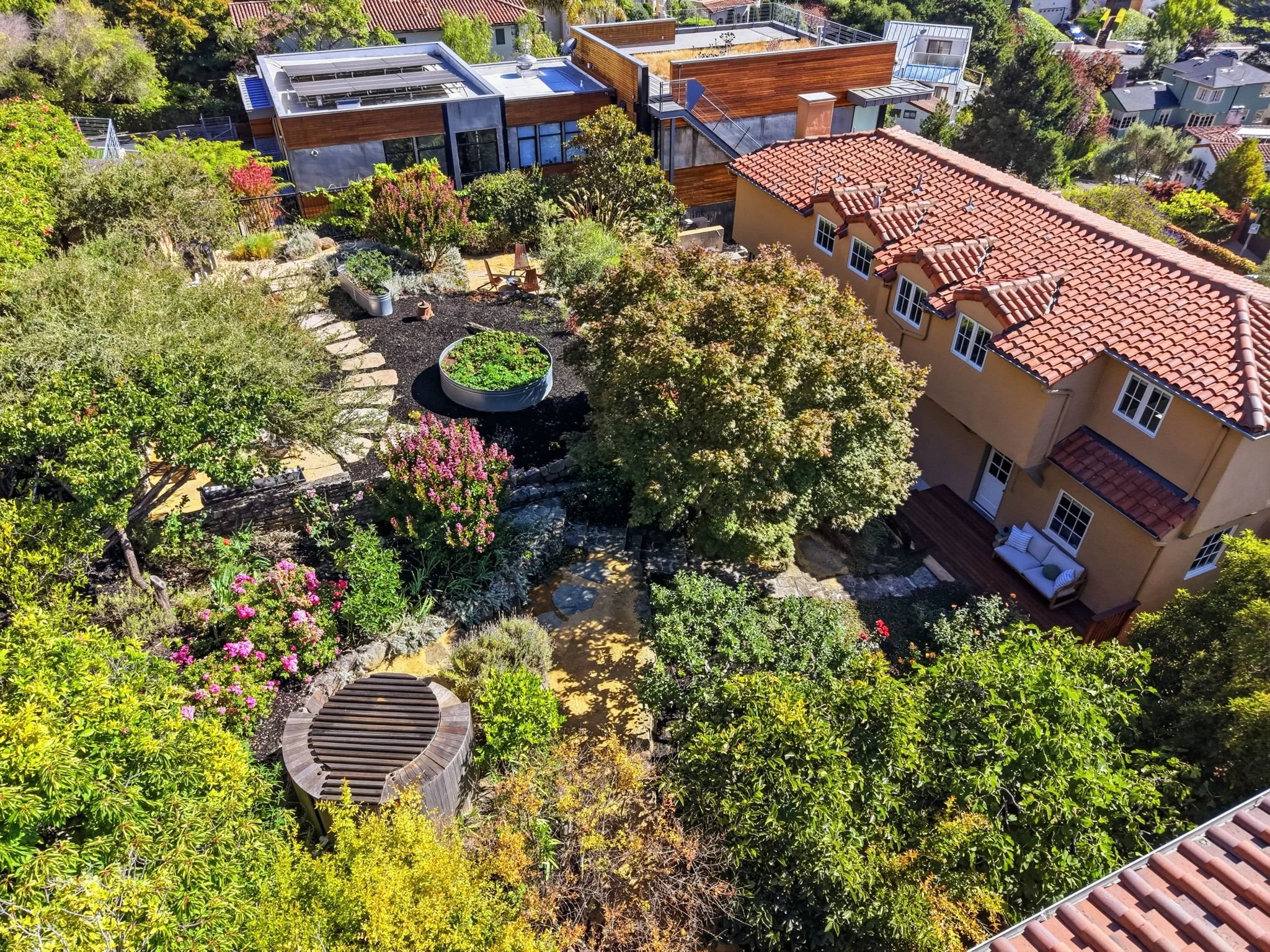 Aerial view of a residential neighborhood with lush trees, colorful plants, and landscaped gardens. There are houses with red-tile roofs and modern buildings with flat roofs, some with solar panels, surrounded by greenery.
