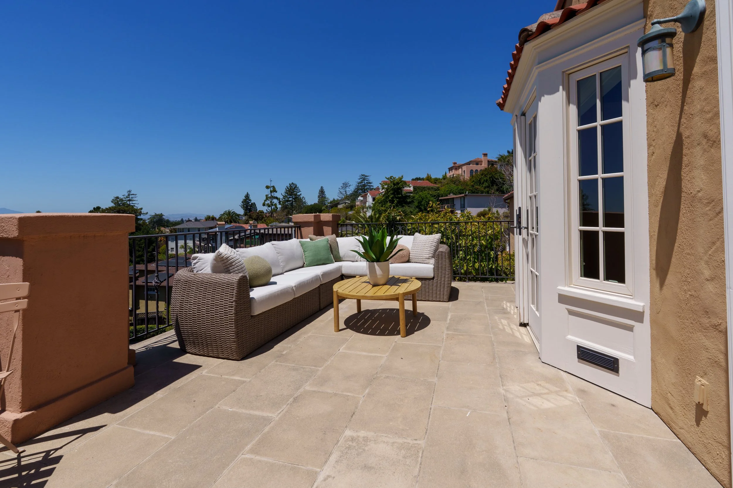 Outdoor patio with beige tiled floor, white sectional sofa with green and beige cushions, round wooden table with potted plant, beige stucco walls, dappled sunlight, clear blue sky, overlooking a residential neighborhood with trees and houses.