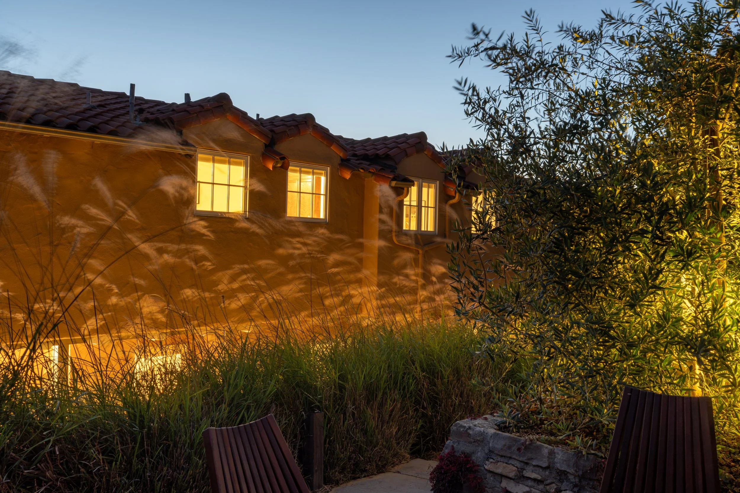 A house with lit windows at dusk, surrounded by greenery and a patio with chairs.