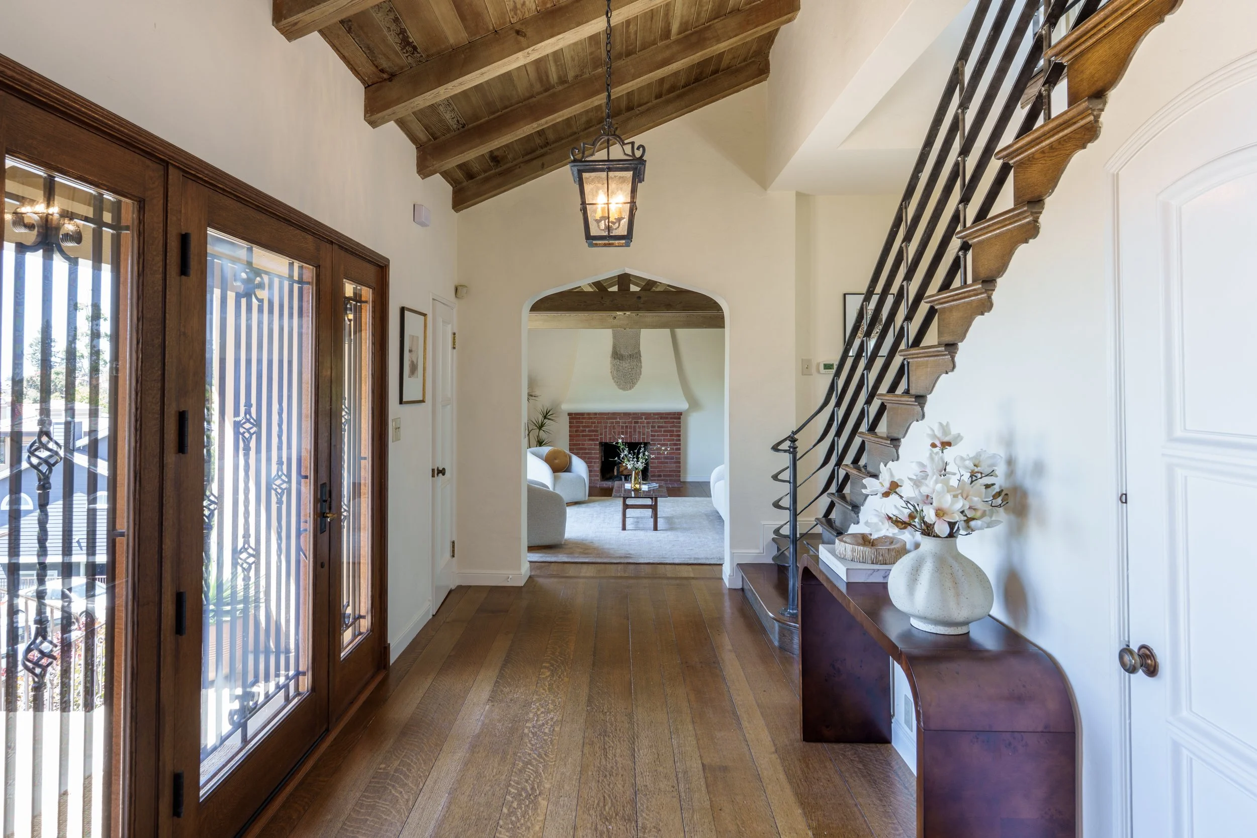 Interior view of a home entryway with wooden flooring, a wooden front door with glass panels, a staircase with wooden steps and black railing, a hanging lantern light, a fireplace with a brick chimney, a white sofa, framed pictures on the wall, and a vase with flowers on a side table.