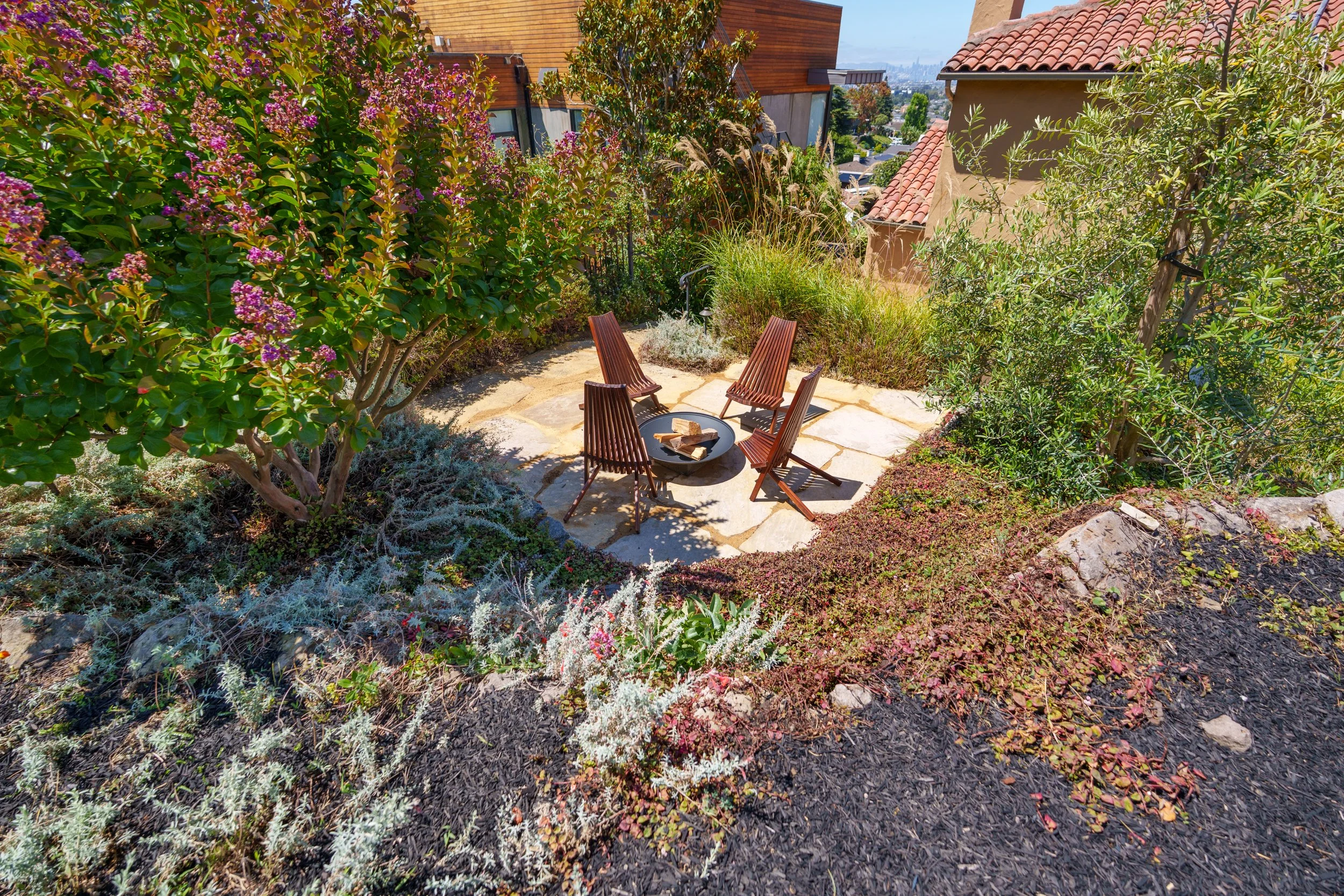 A backyard patio surrounded by lush greenery, including flowering bushes and small trees, with four wooden chairs arranged around a fire pit, in a residential neighborhood with neighboring houses and rooftops visible in the background.