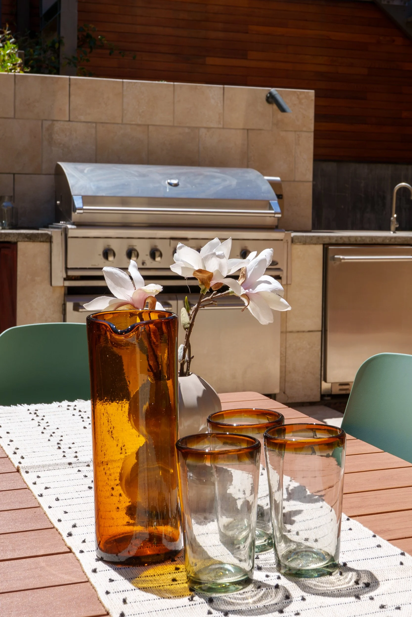 Outdoor patio table with decorative amber-colored glass vases, a white flowers in a vase, and four empty glass tumblers. In the background, an outdoor BBQ grill and a beige tile wall with a wall-mounted light fixture.
