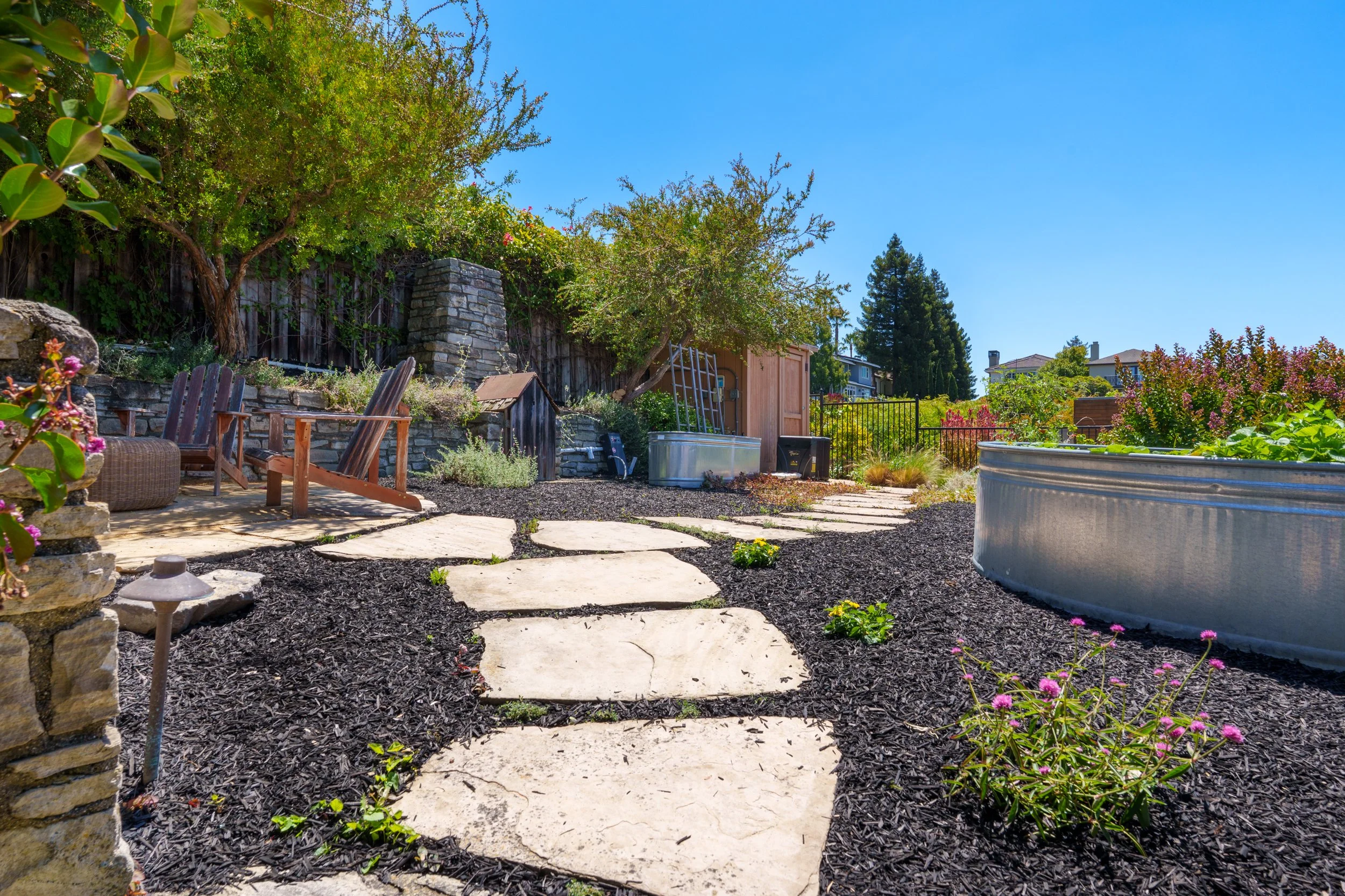 A backyard garden with a stone pathway, potted plants, outdoor chairs, a small shed, and lush greenery under a clear blue sky.