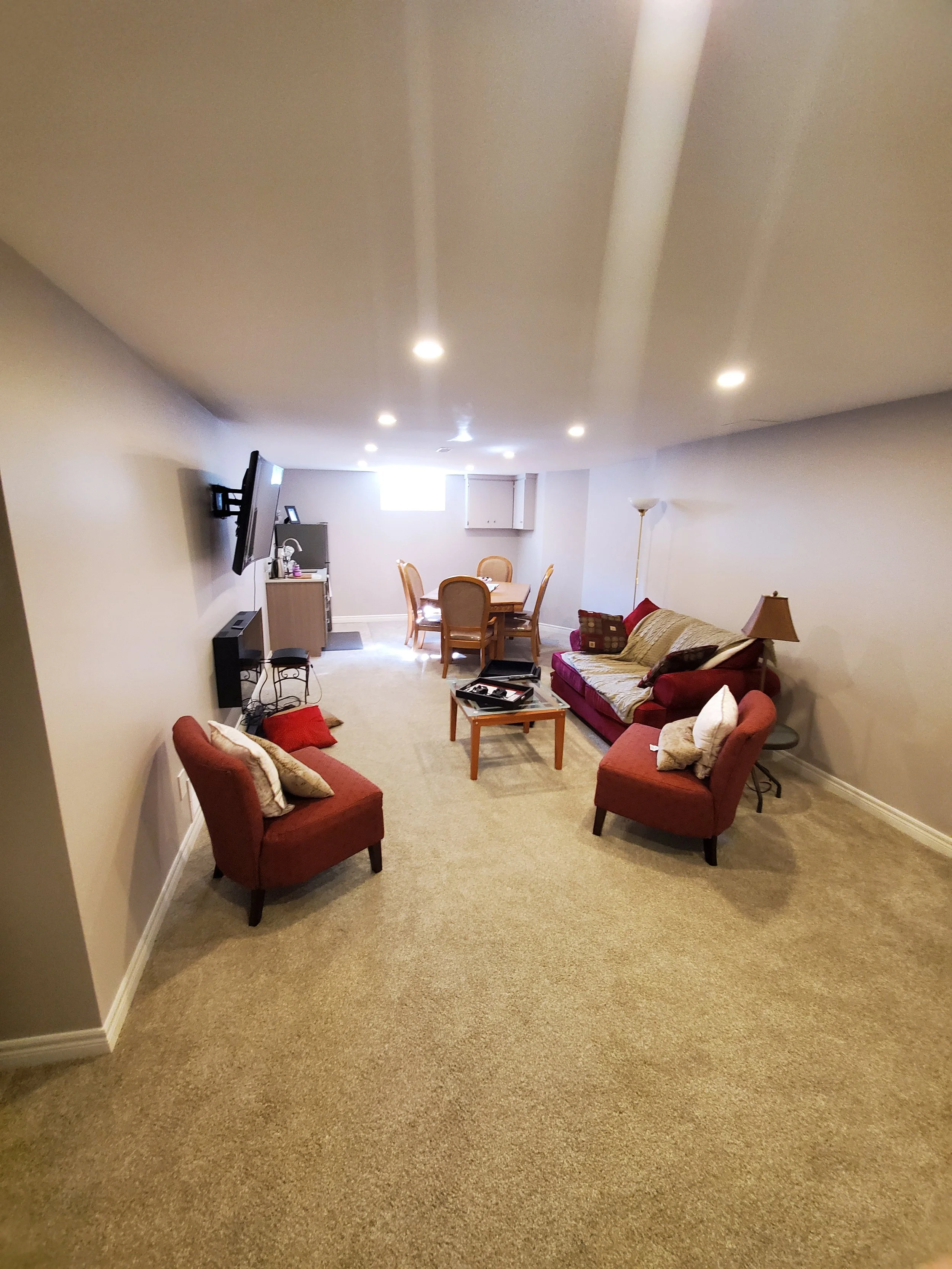 A spacious living room with beige carpet, three red chairs with throw pillows, a wooden coffee table, a sofa with pillows, a floor lamp, a wall-mounted TV, a small table with items, and a dining table with four chairs near a small window.