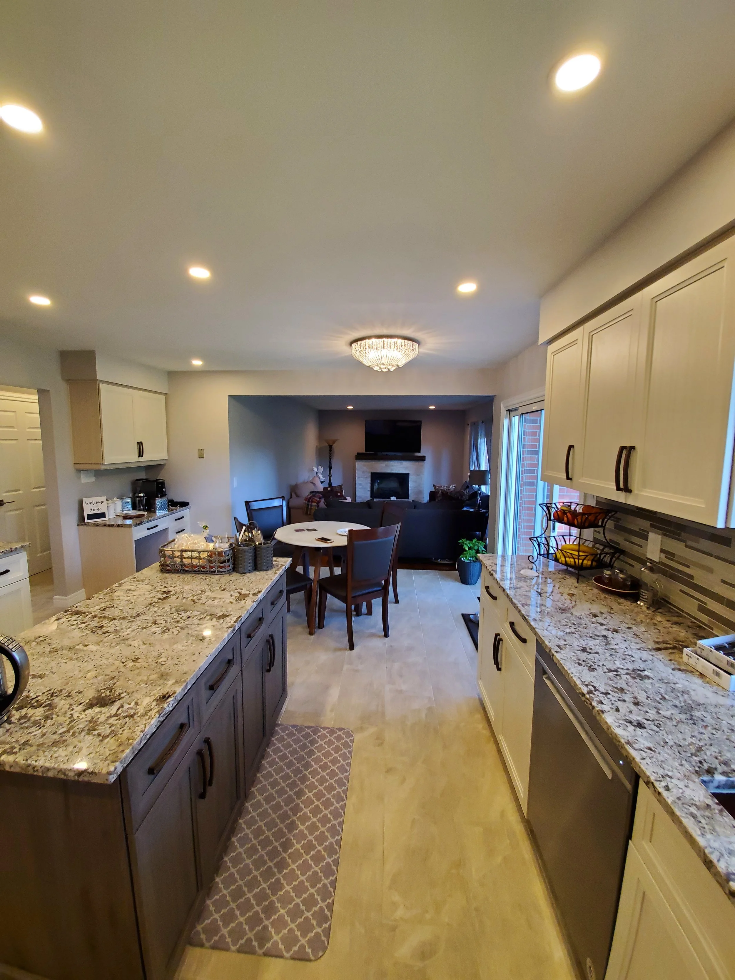 A spacious open kitchen with granite countertops, dark lower cabinets, white upper cabinets, and a dining area in the background with a living room and fireplace.