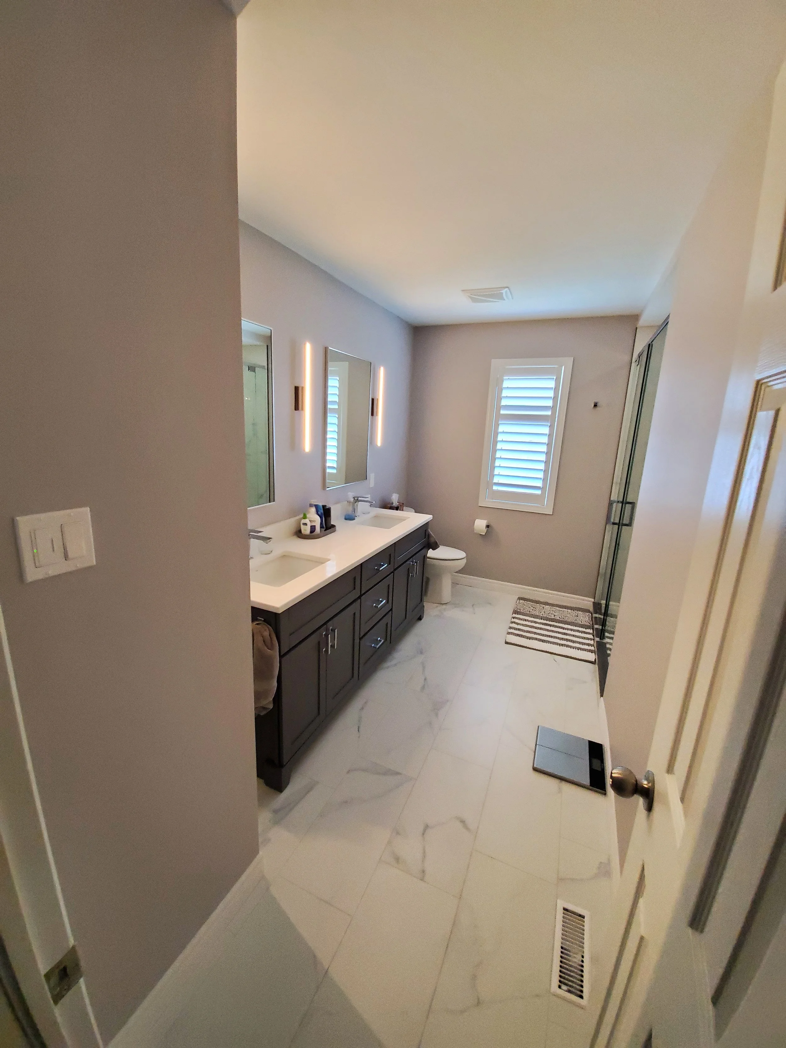 A modern bathroom with gray walls, white marble floor tiles, a double sink vanity with dark cabinets, two mirrors, and wall-mounted lights. There is a window with shutters, a toilet, a small striped rug, and a digital scale on the floor.