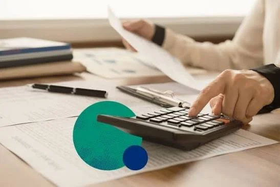 Person using a calculator and holding papers at a desk with pens and documents.