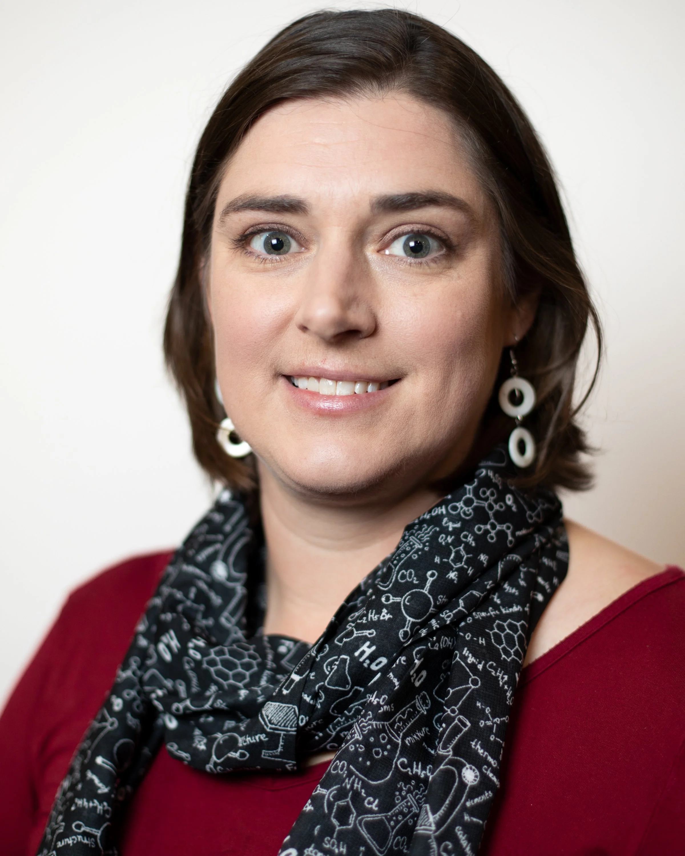 Close-up of a smiling woman with shoulder-length brown hair, wearing a black scarf with scientific chemical formulas and a maroon top, against a plain white background.