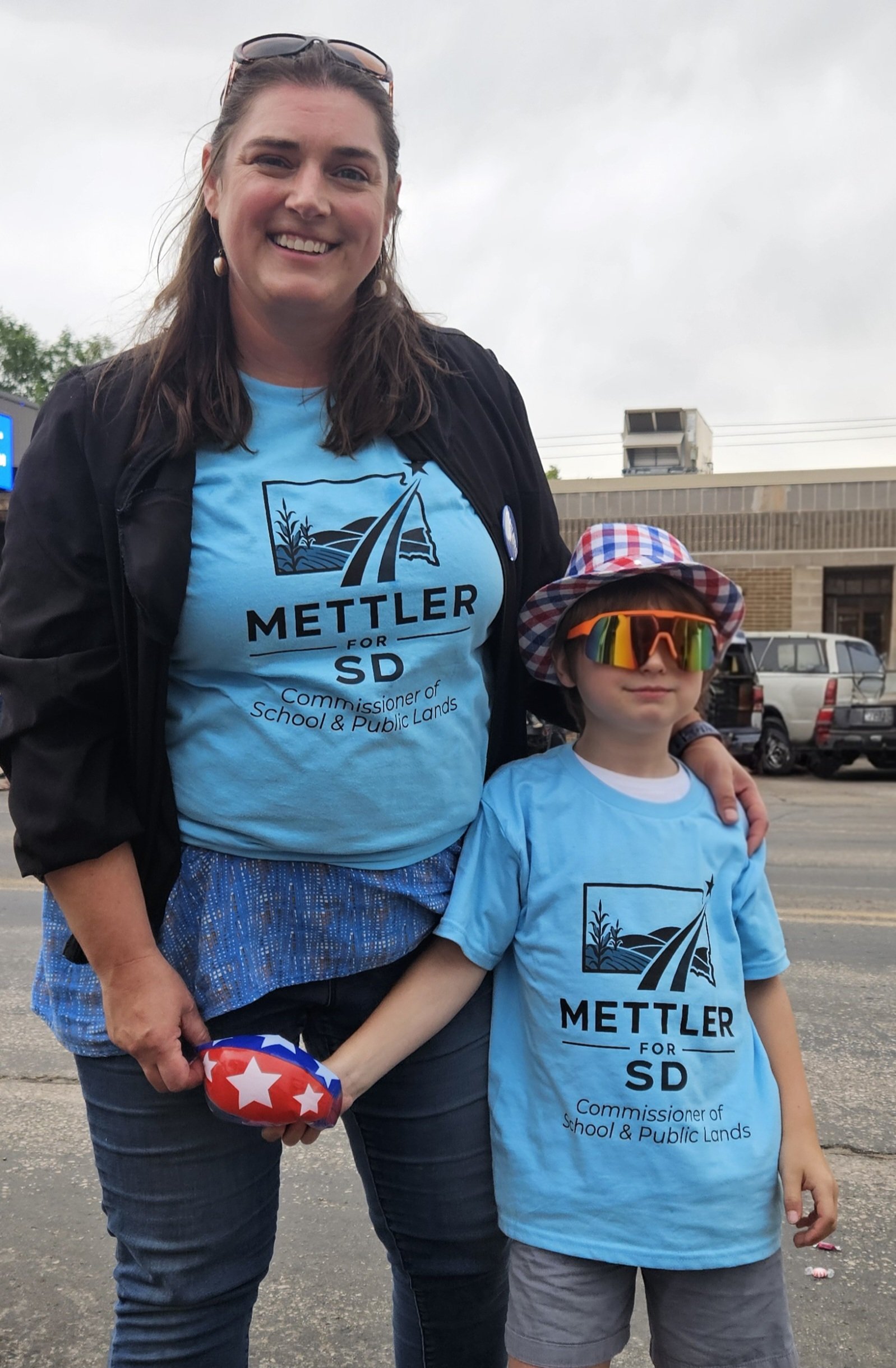 A woman and a young boy standing outdoors in a parking lot. Both are wearing light blue T-shirts with a logo that says 'METTLER FOR SD' and additional text. The woman has dark brown hair, sunglasses on her head, and is smiling. The boy is wearing sunglasses, a checkered hat, and holding a small American flag. Overcast sky and some parked cars are visible in the background.