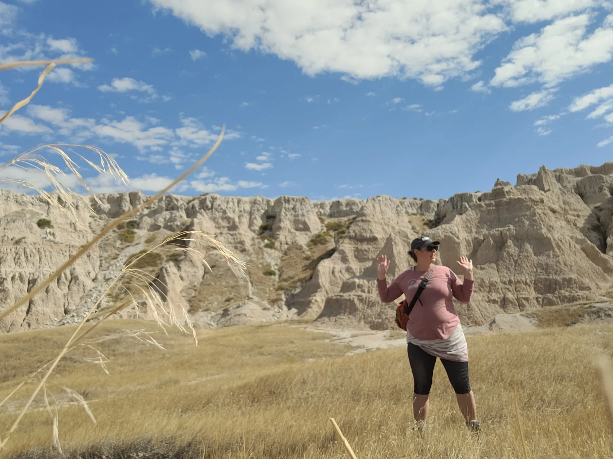 A woman wearing sunglasses, a pink shirt, black pants, and a backpack walking outdoors near rocky formations under a blue sky with white clouds.