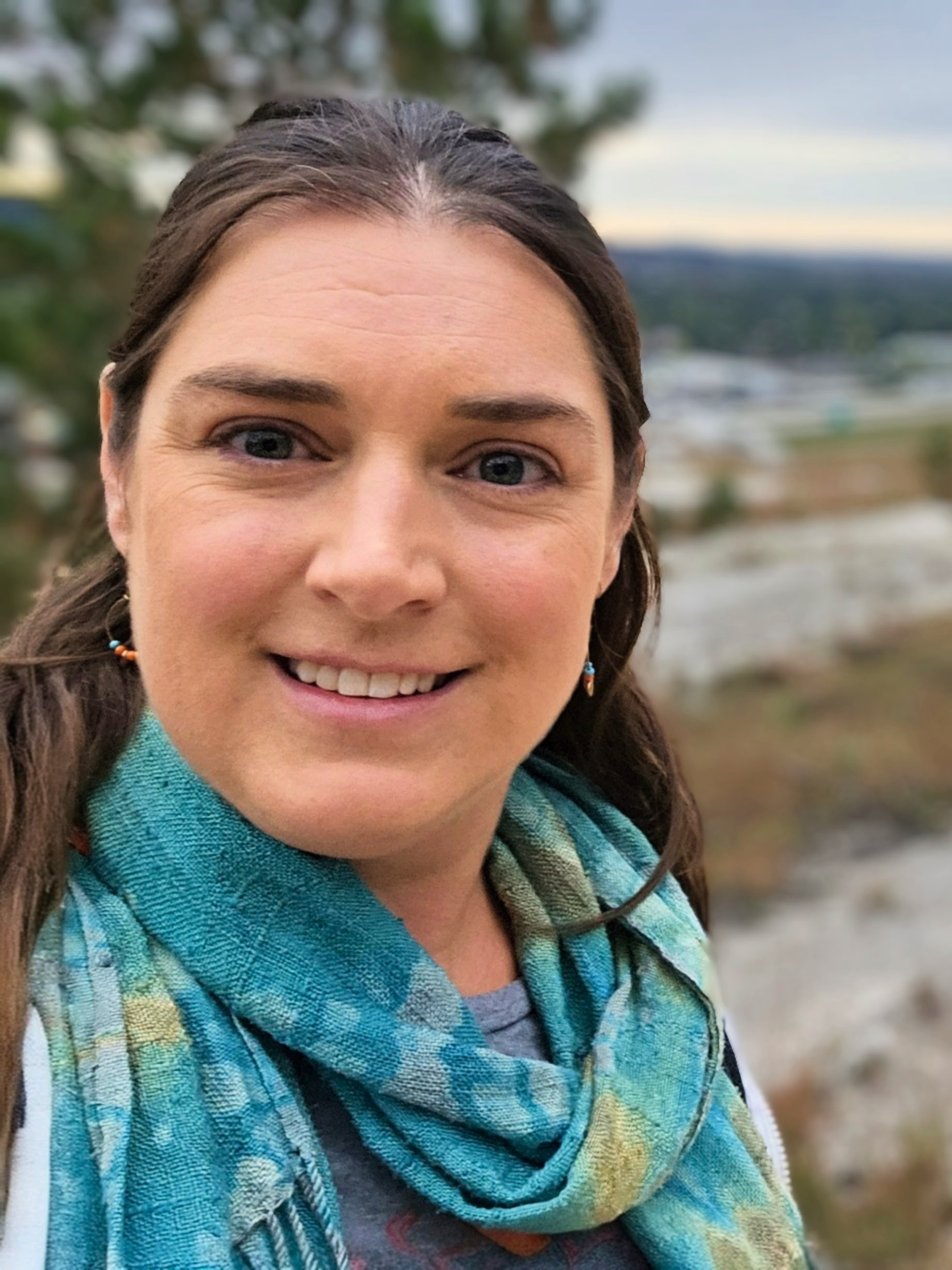 Close-up of a woman with brown hair, blue eyes, smiling, outdoors with trees and a body of water in the background.