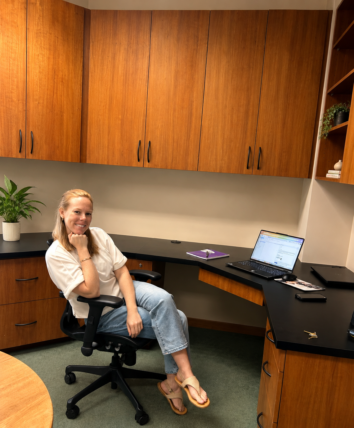A woman sitting in an office chair in a small office, smiling at the camera, with a desk, closed laptop, purple notebook, and keys on the desk, and a potted plant on the side.