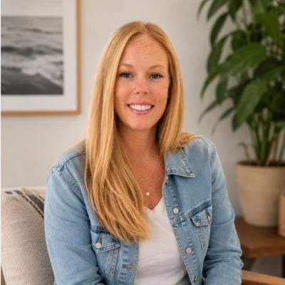 A young woman with long red hair, wearing a denim jacket and white shirt, smiling while sitting in a cozy room with a plant and framed picture in the background.