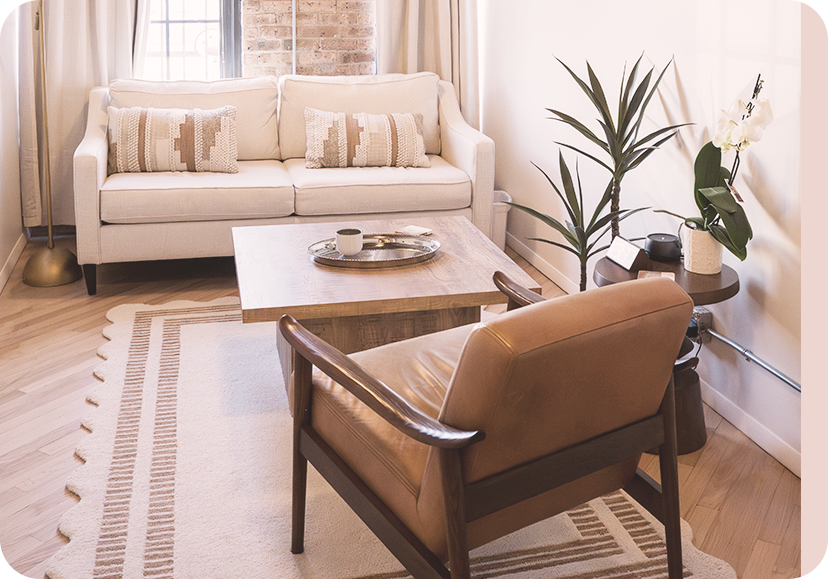 Living room with beige sofa, wooden coffee table, tan armchair, and potted plants near a window with light curtains.