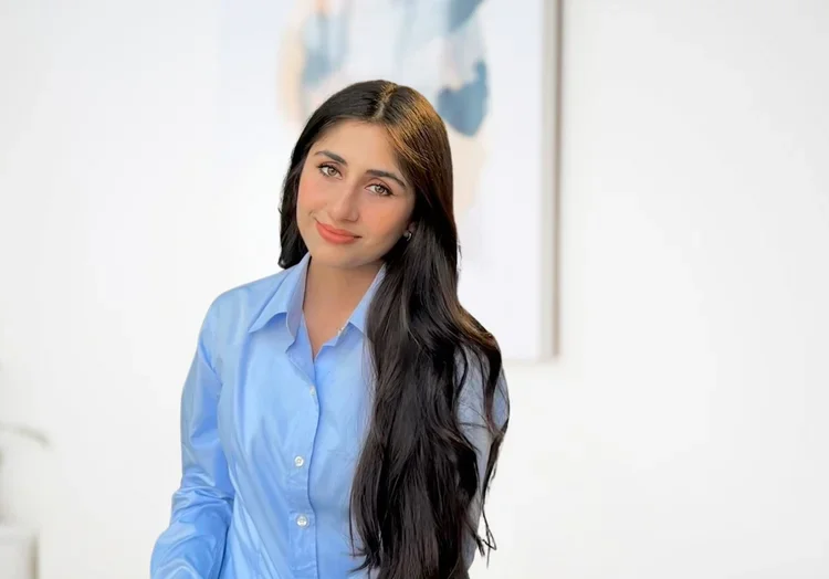 Young woman with long dark hair wearing a light blue button-up shirt, smiling in an office setting.
