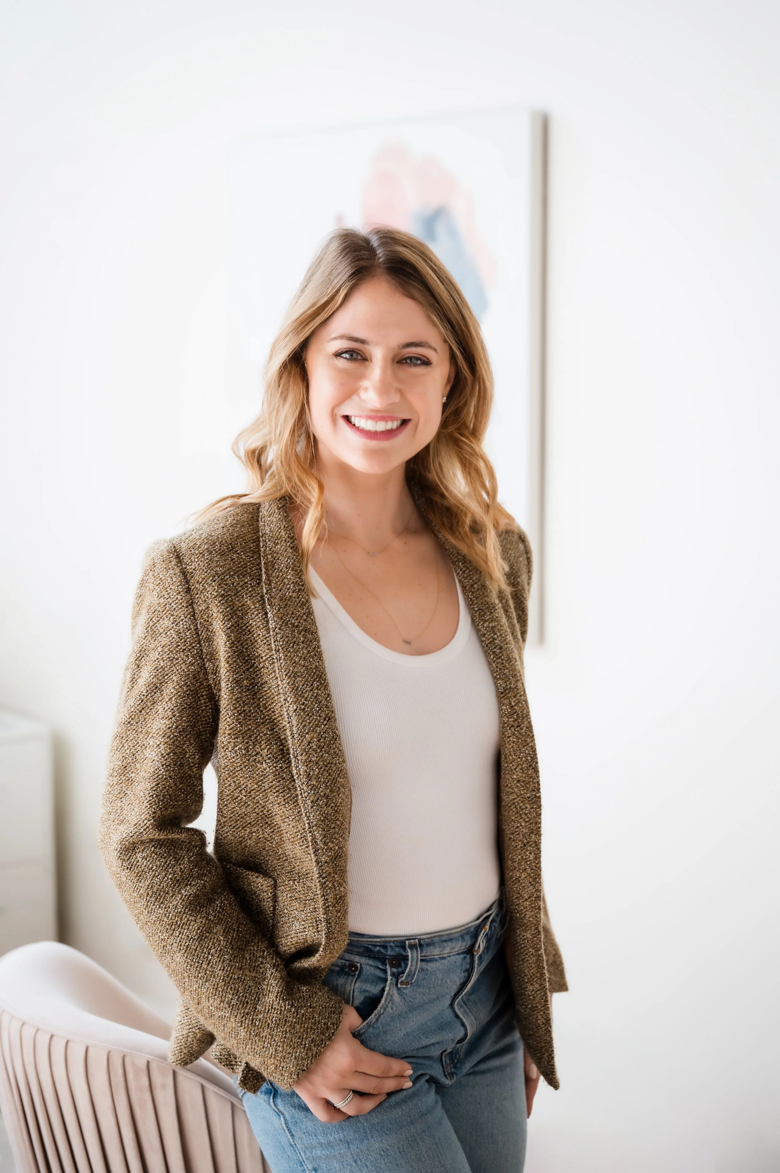 A woman with shoulder-length blonde hair smiling, wearing a brown blazer over a white top, with blue jeans, standing in a bright, modern room with a blurred artwork on the wall in the background.
