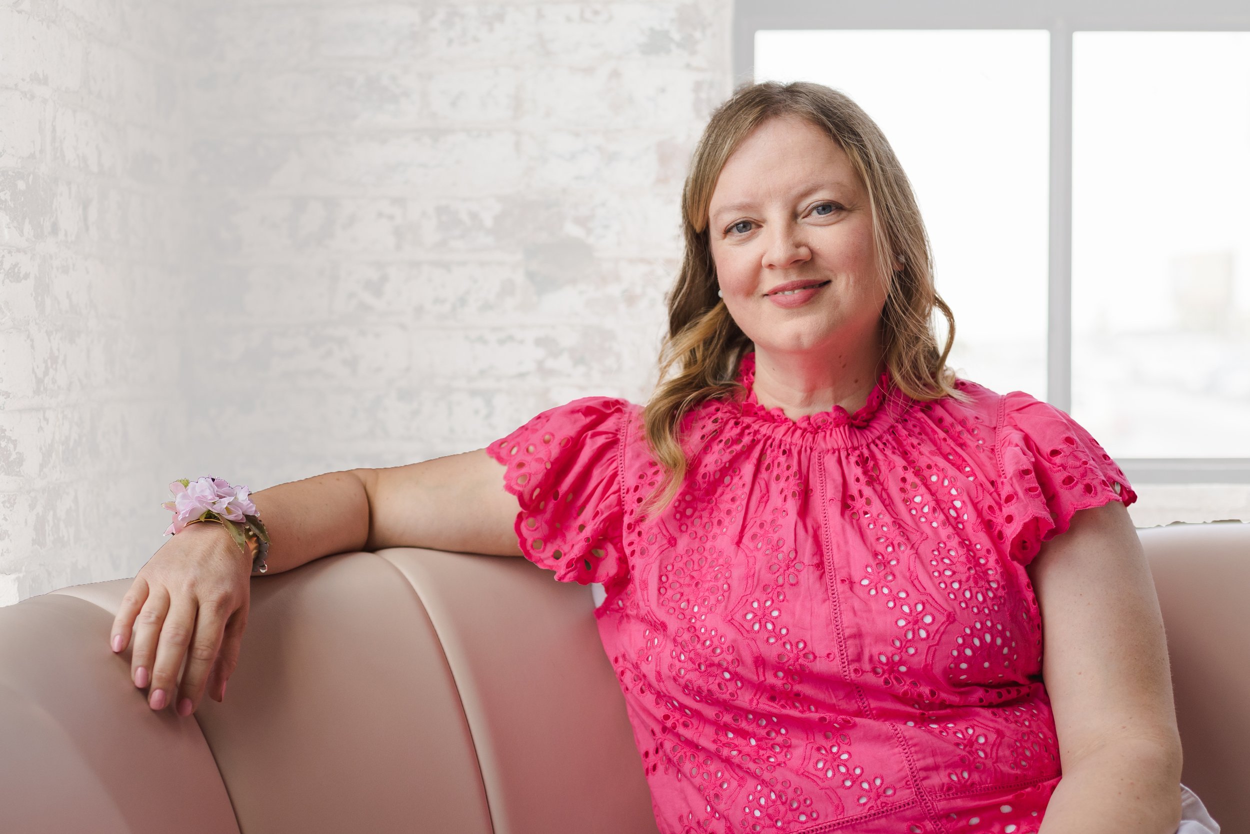 A woman with light skin and blonde hair wearing a bright pink blouse with eyelet details, sitting on a light-colored couch in a room with white brick walls and bright windows, smiling at the camera.
