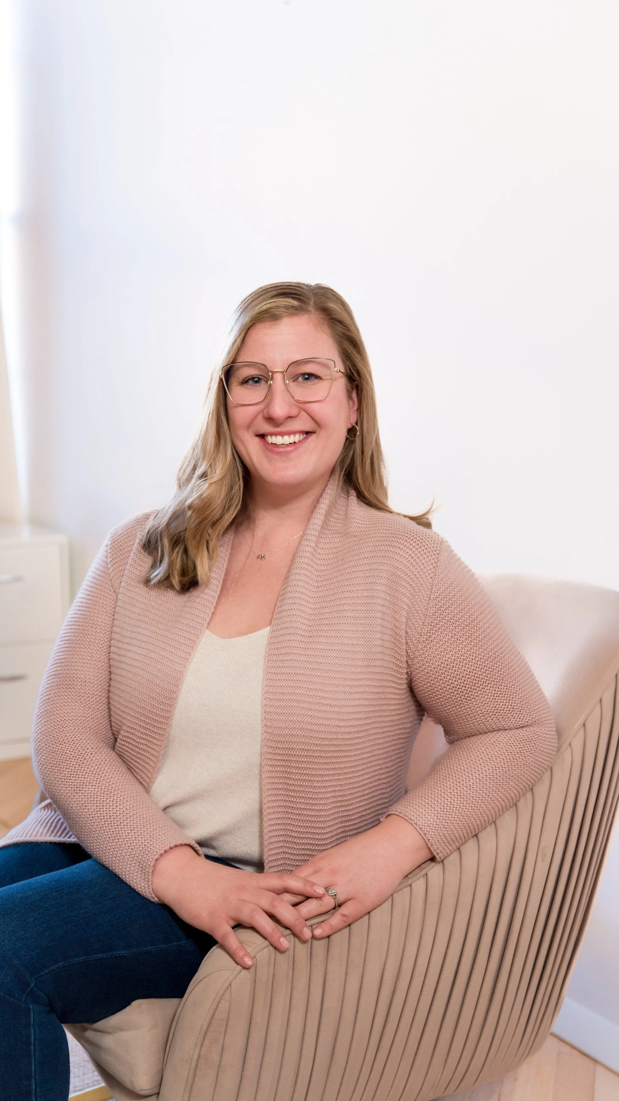 A woman with blonde hair, glasses, and a warm smile sitting on a beige striped armchair in a room with white walls and light wood flooring.