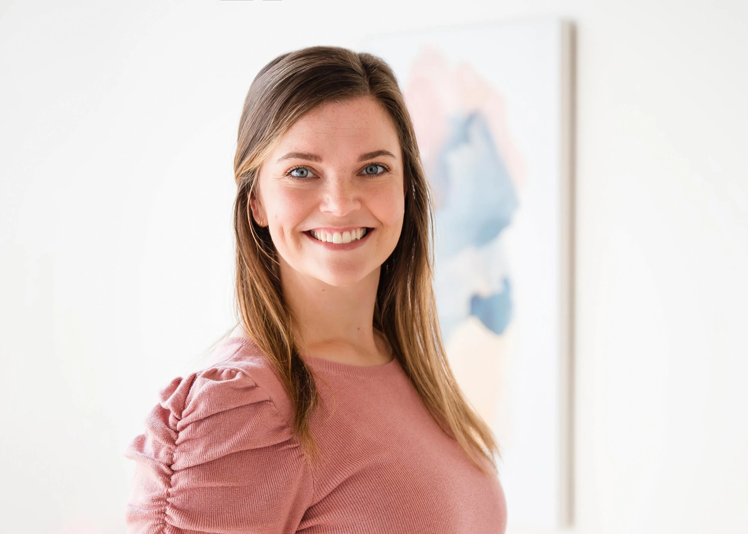 A woman with long brown hair wearing a pink top, smiling and looking at the camera, standing in front of a white wall with a painting in the background.
