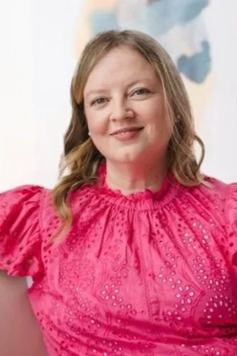 A woman with shoulder-length light brown hair wearing a bright pink embroidered blouse, smiling in front of a colorful abstract background.