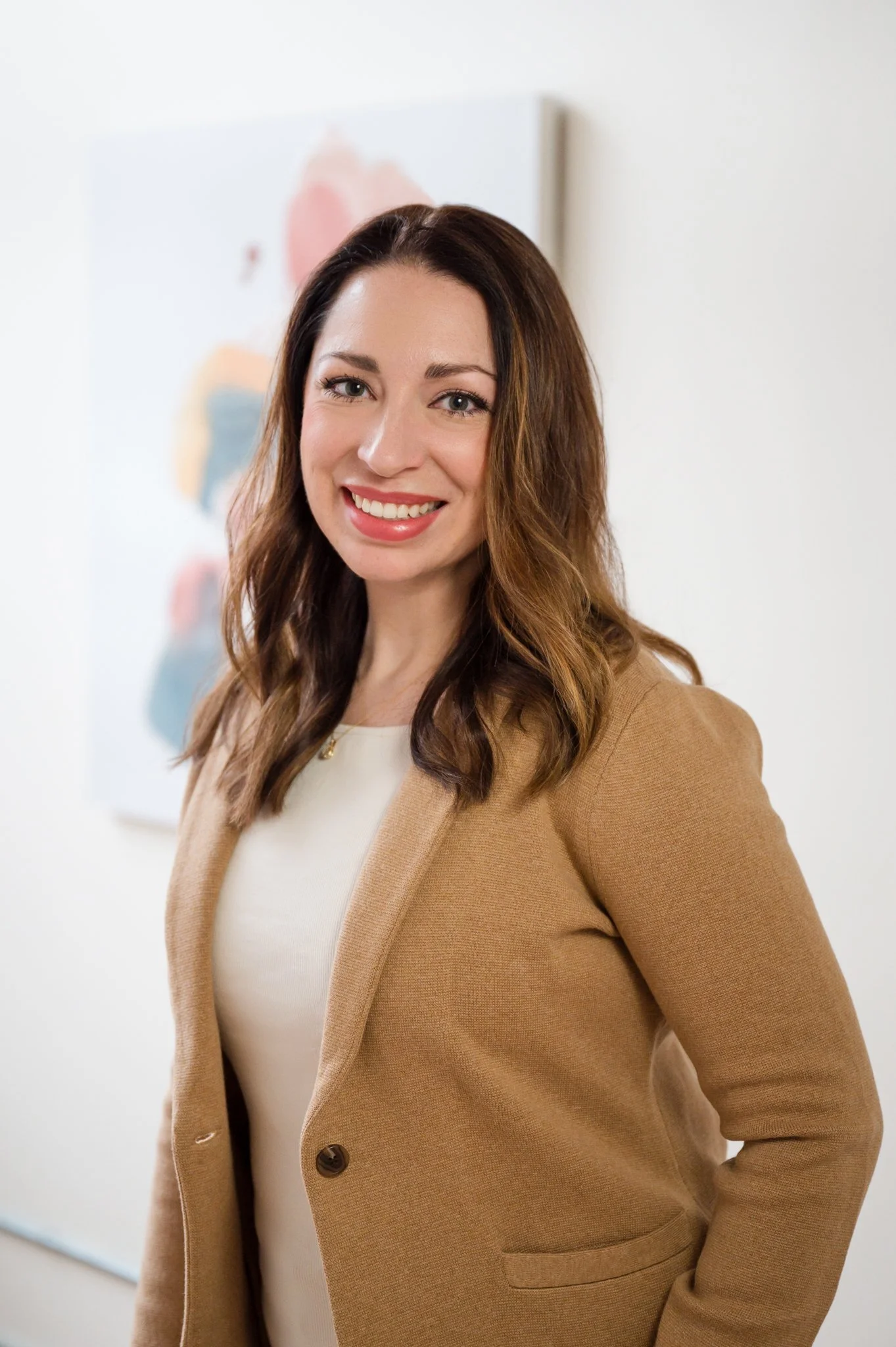 A woman with long brown hair, wearing a beige blazer over a cream blouse, smiling at the camera in an indoor setting with a blurry colorful abstract painting in the background.