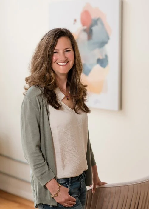 A woman with long brown hair smiling and standing indoors with abstract artwork in the background.