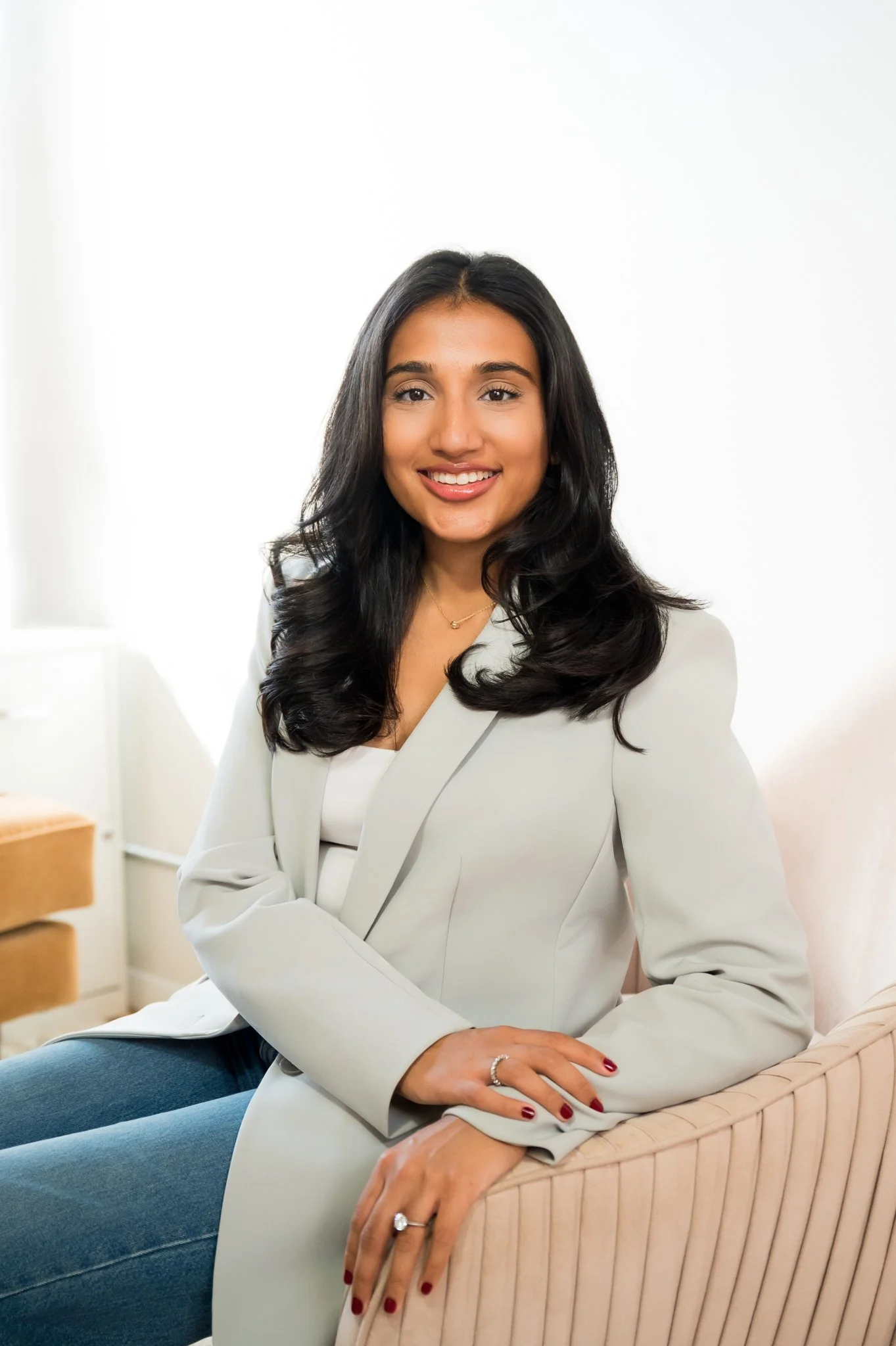 A woman with dark, wavy hair and tan skin, smiling and wearing a white blazer, gold necklace, and rings, sitting on a beige cushioned chair in a bright, minimalistic room with white walls.
