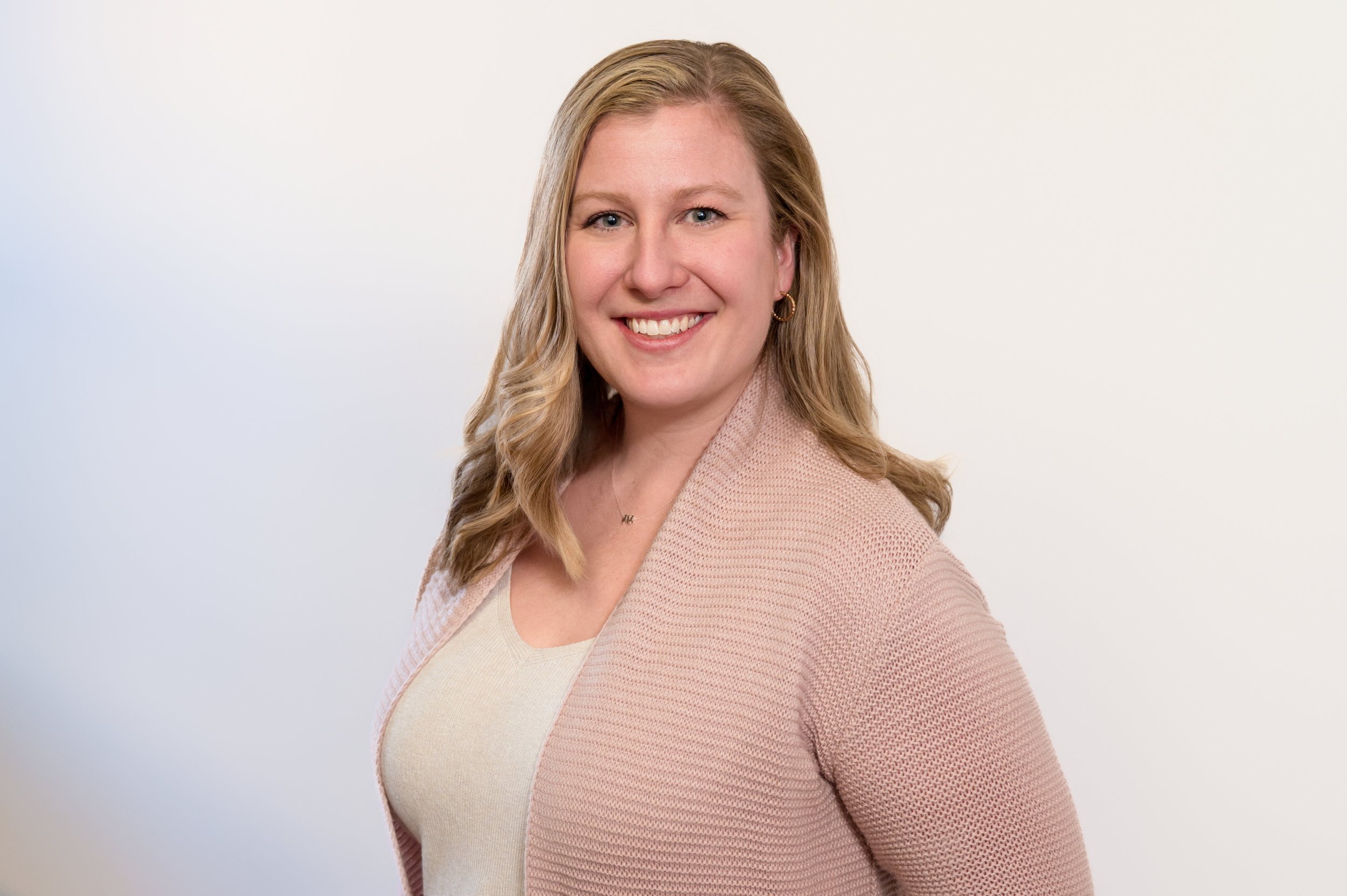 Portrait of a smiling woman with blonde hair, wearing a pink and cream sweater, posing against a plain white background.