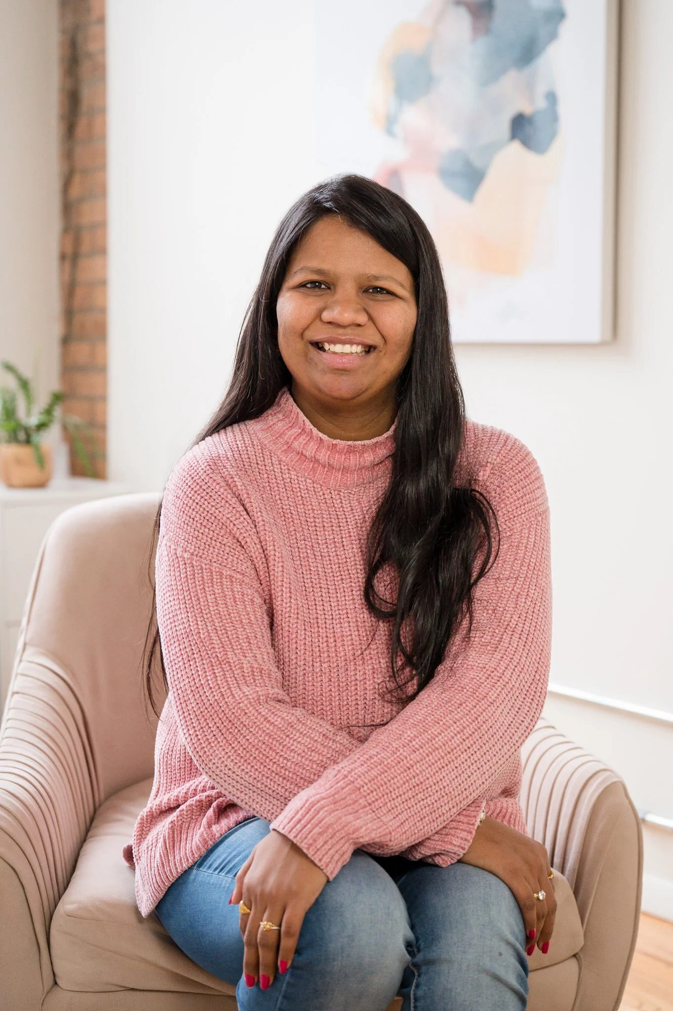 A woman with long dark hair smiling, seated on a beige armchair in a well-lit room, wearing a pink knit sweater and blue jeans, with artwork on the wall behind her and a small potted plant in the background.